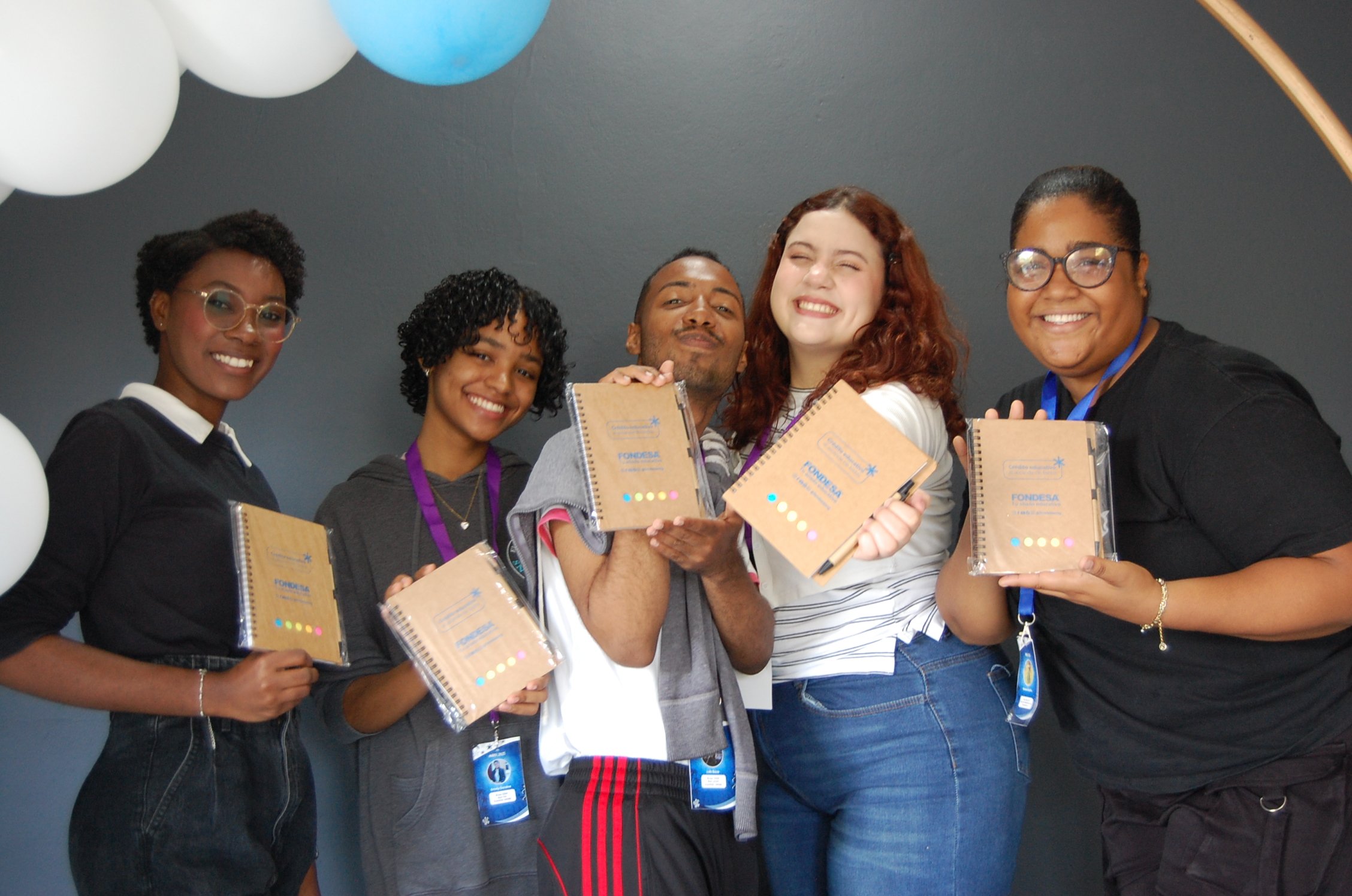 Grupo de cinco personas jóvenes sonriendo y sosteniendo cuadernos, en un evento o ceremonia, decoraciones con globos blancos y azules en el fondo.