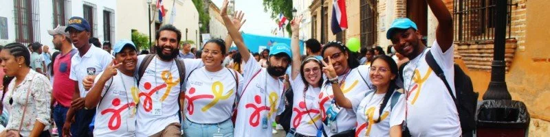 Grupo de personas participando en un desfile o manifestación en una calle, algunos con camisetas blancas con un lazo amarillo impreso, levantando las manos y sonriendo.