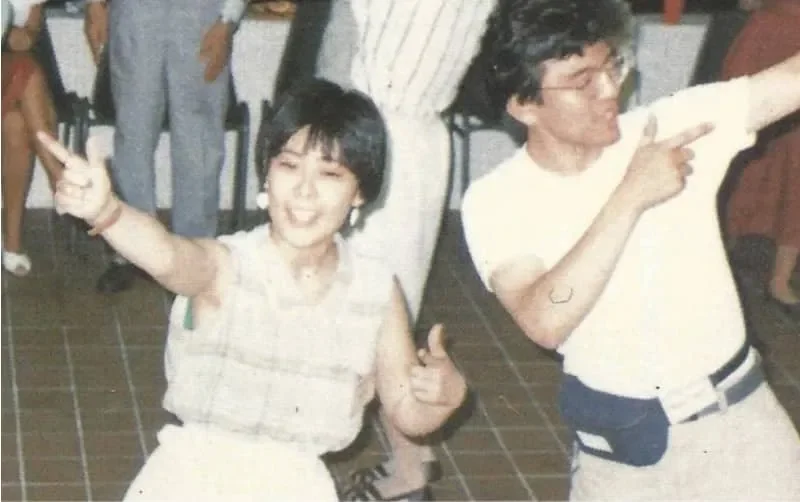 Dos personas bailando y posando alegremente en una fiesta o evento social, con varias personas en el fondo.