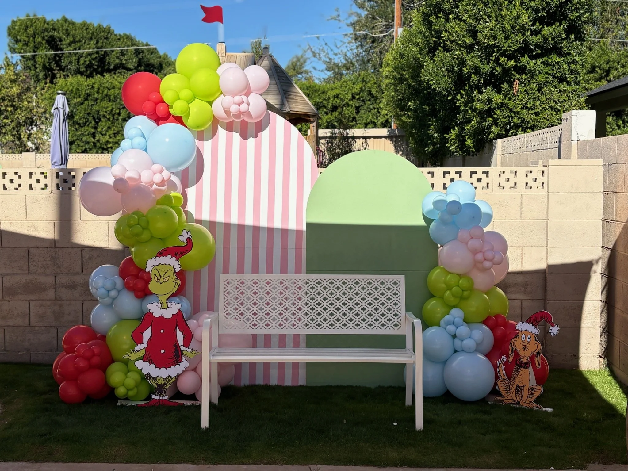 Festive outdoor Christmas scene with balloon arrangements, cartoon Christmas characters, and a white bench in front of a cream brick wall, with trees and a blue sky in the background.