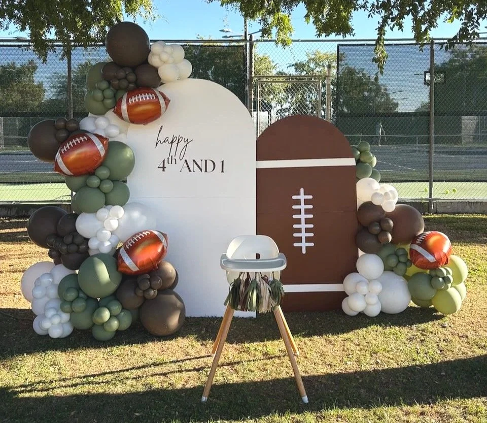 Balloon arch with football-themed decorations against a fence on a grassy field, with a high chair in front and a football-shaped backdrop for a celebration.