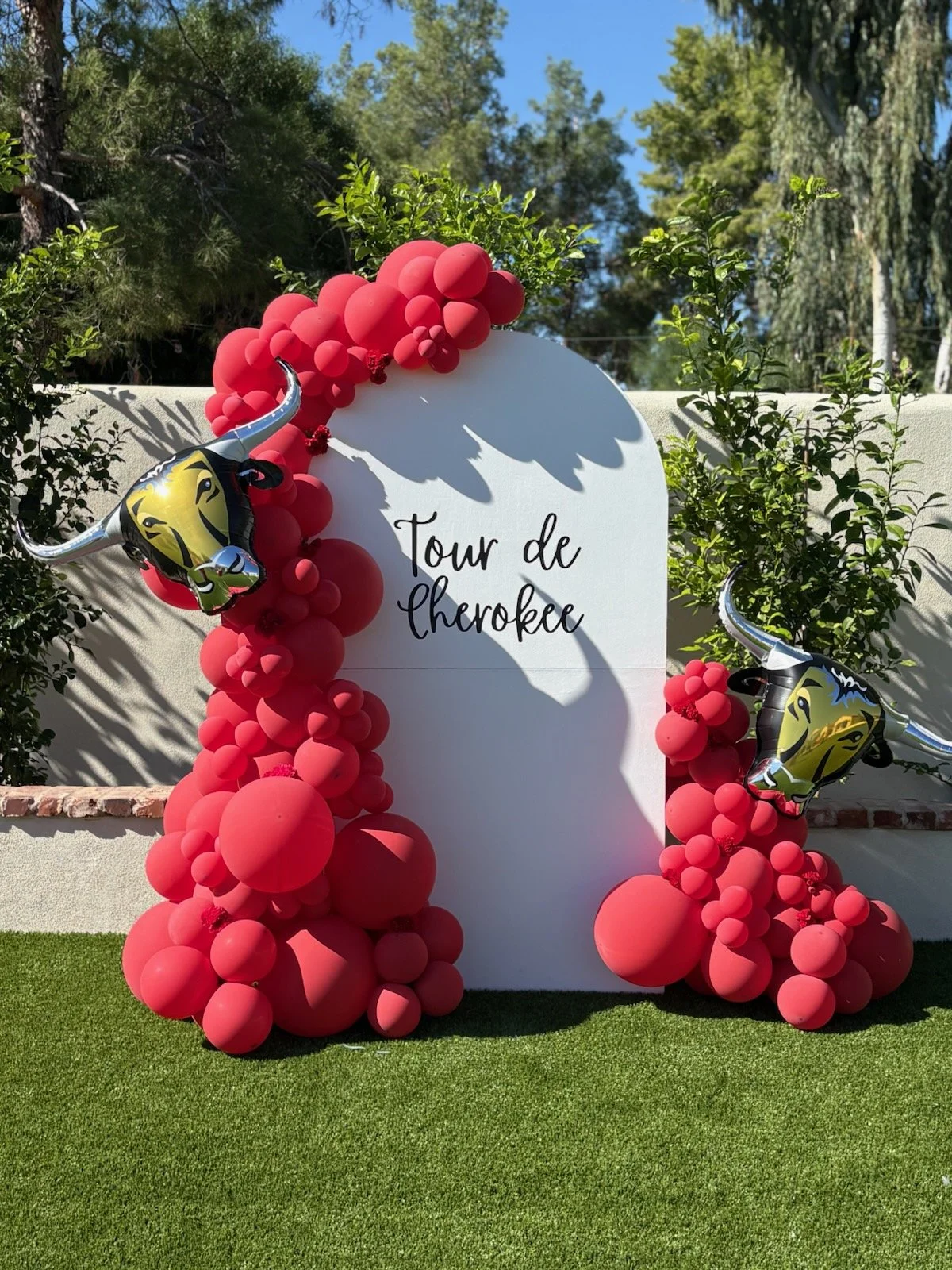 Decorative balloon arrangement with red balloons surrounding a white sign reading 'Tour de Cherokee' in black script, flanked by two balloons shaped like a longhorn steer head with a yellow face and black markings. The setup is outdoors on a lawn wit