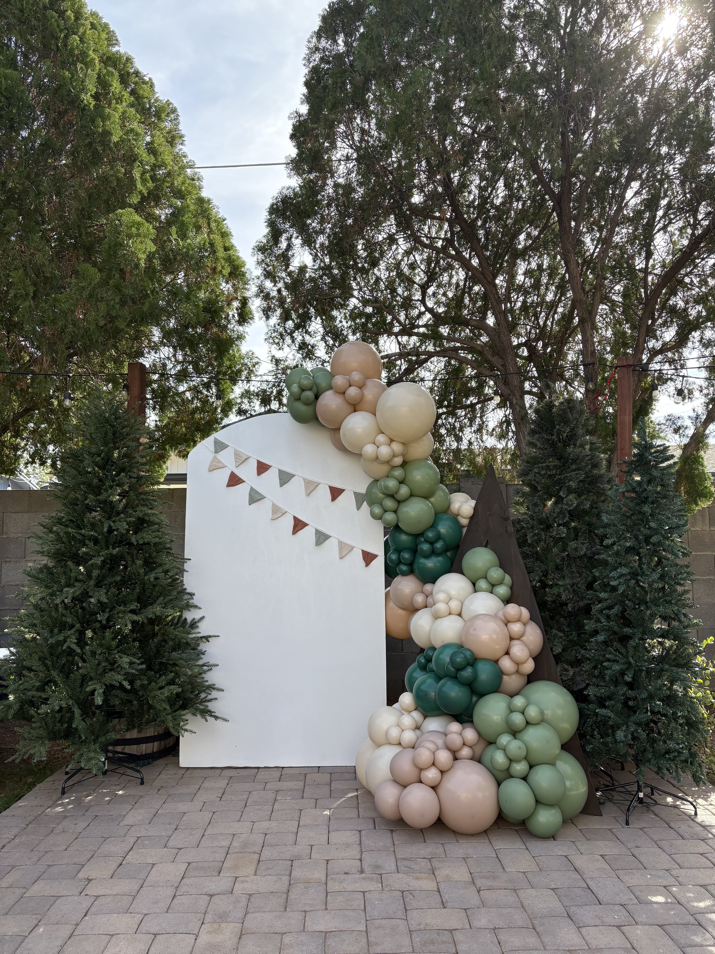 Decorative outdoor setup with a white board, green and white balloons, bunting, two small evergreen trees, and a large tree in the background.