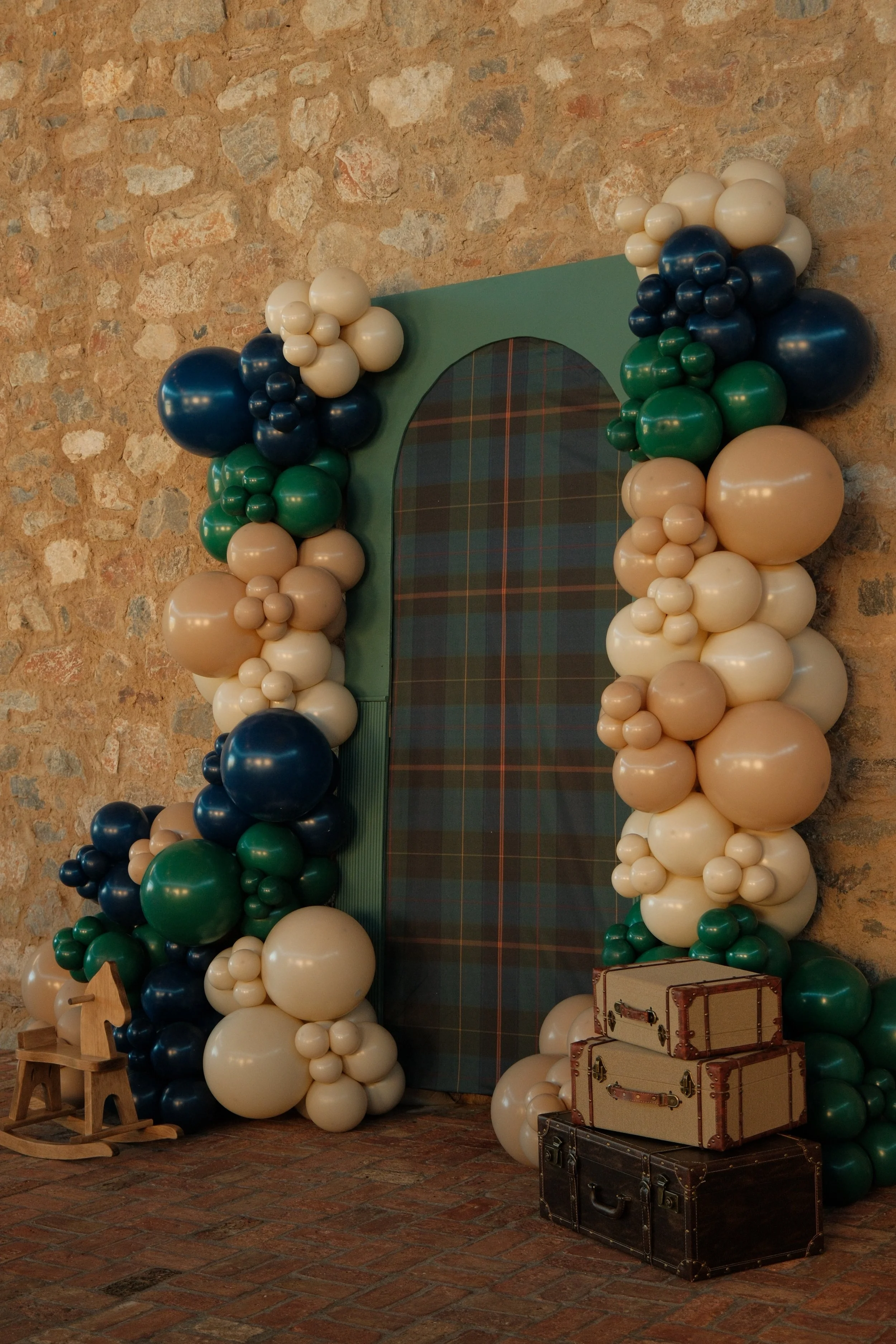 A wall decorated with a large arch-shaped mirror surrounded by clusters of balloons in blue, green, beige, and white colors, with a small wooden rocking horse and stacked vintage suitcases on the floor in front of the wall.