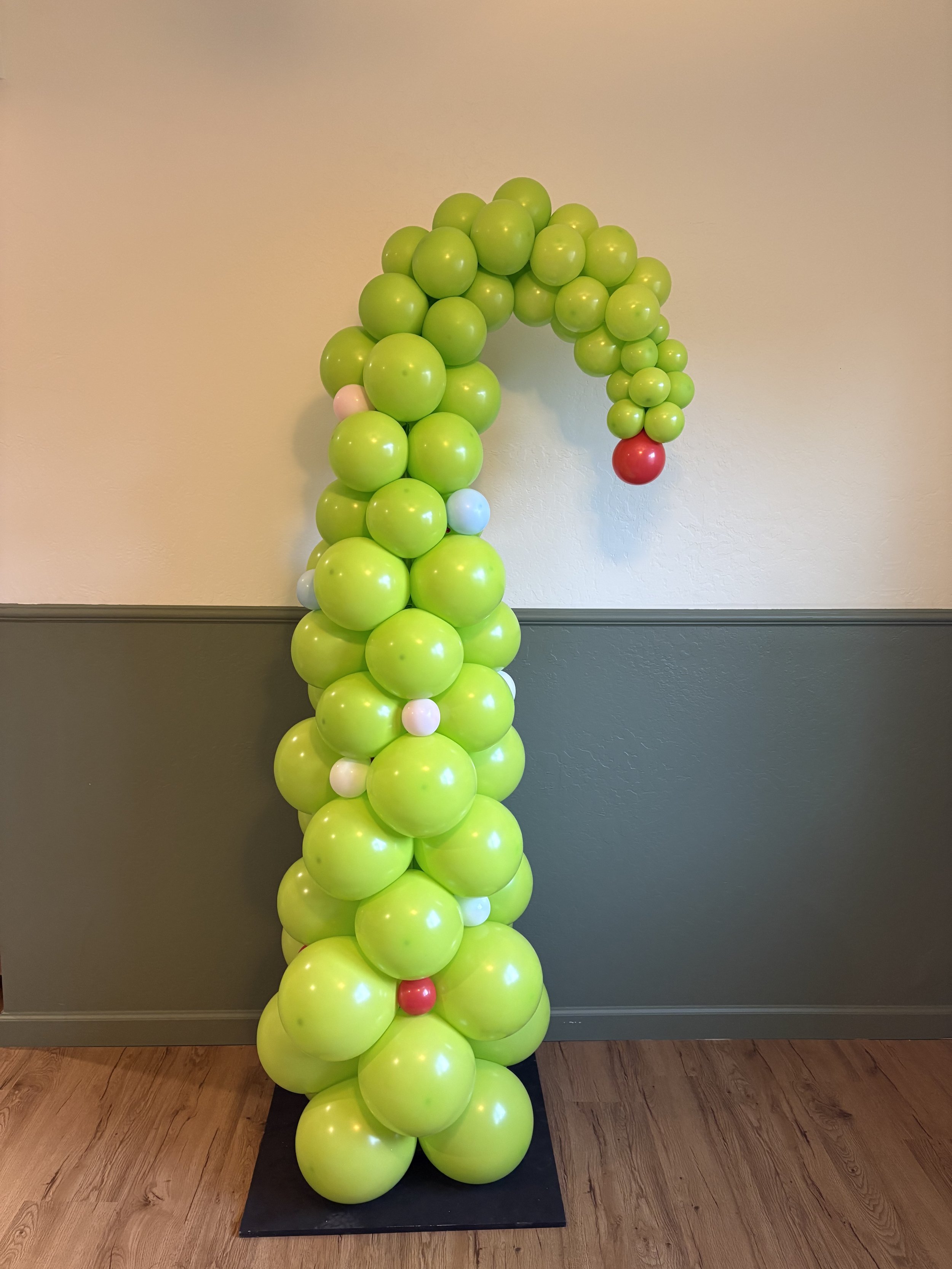 A candy cane-shaped balloon sculpture made of green balloons with red and white accents, standing on a black base indoors.