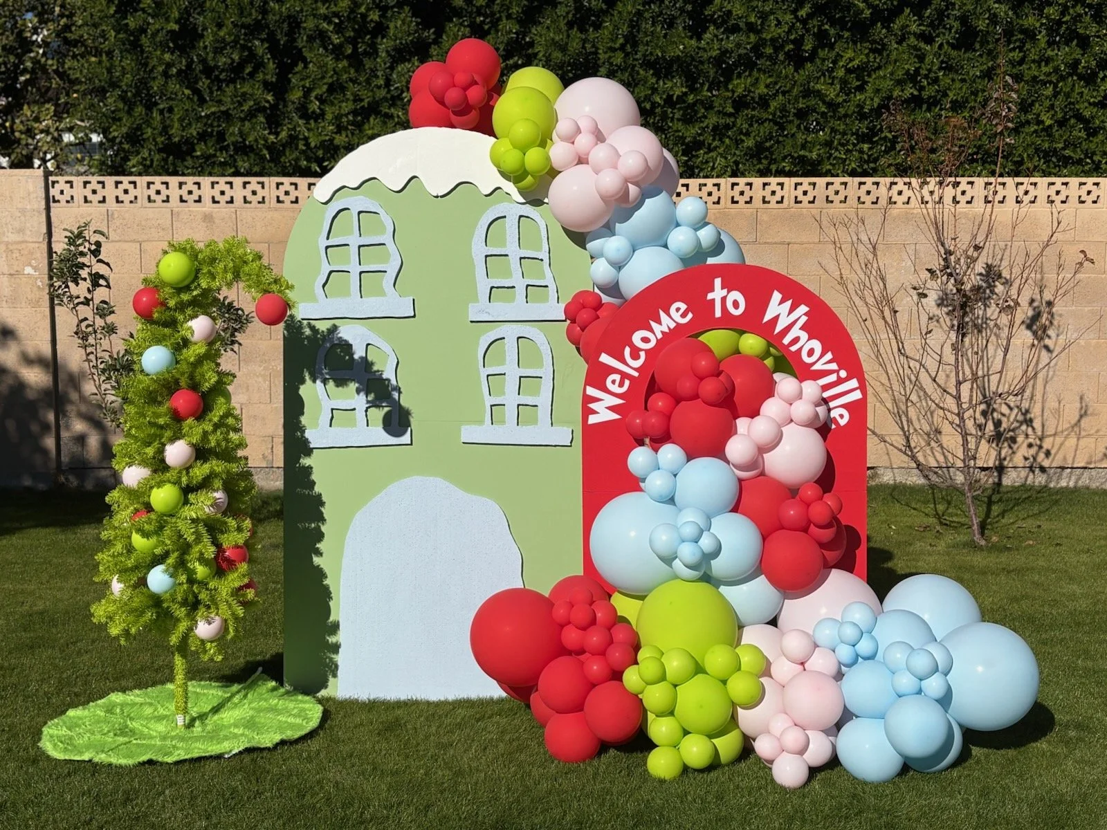Colorful holiday display with a small green house, a decorated Christmas tree, and a red arch with the text "Welcome to Whoville" surrounded by red, white, pink, blue, and green balloons on a grassy lawn.