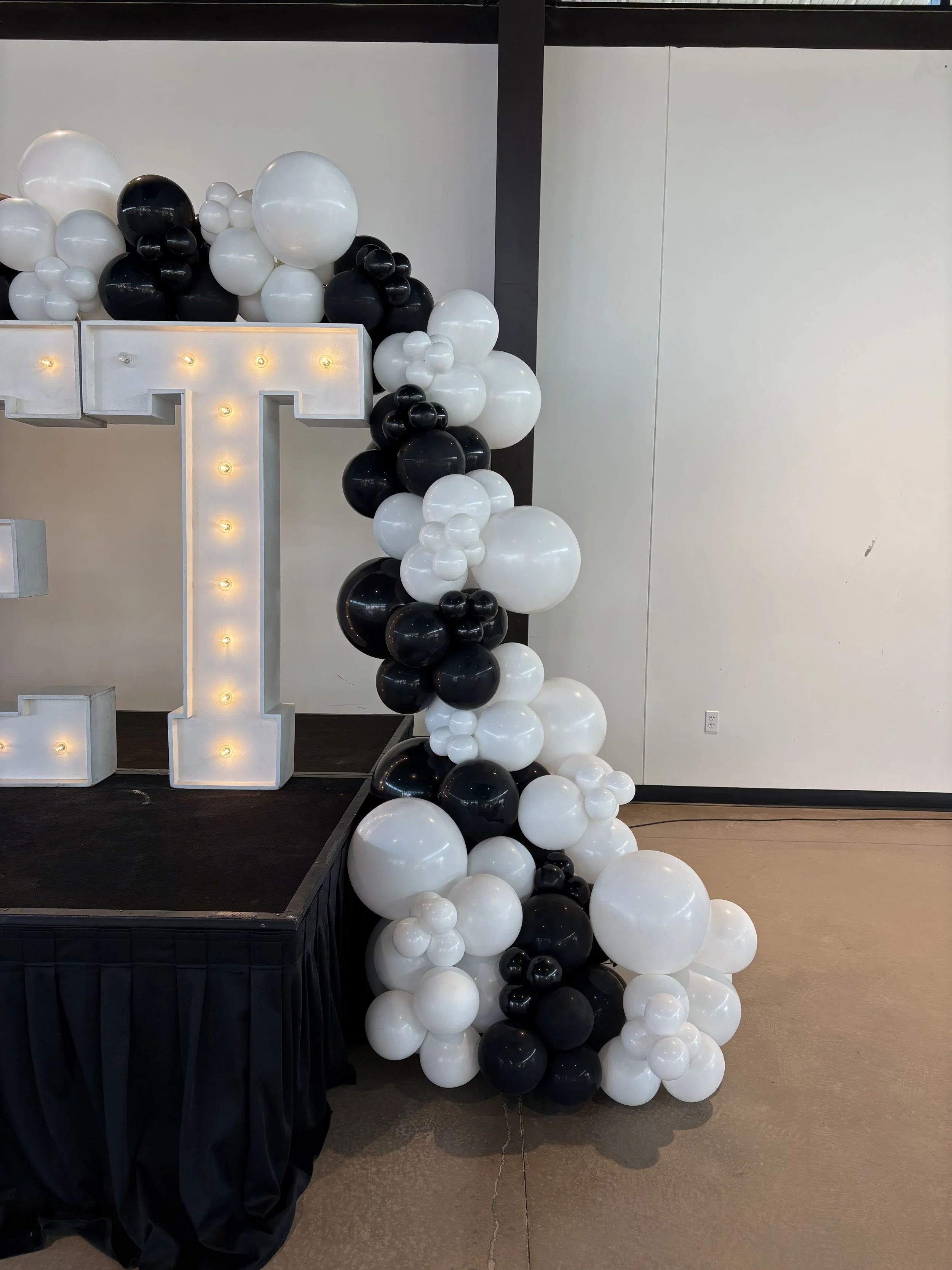 Decorative display with black and white balloons arranged around a large illuminated letter T on a stage.