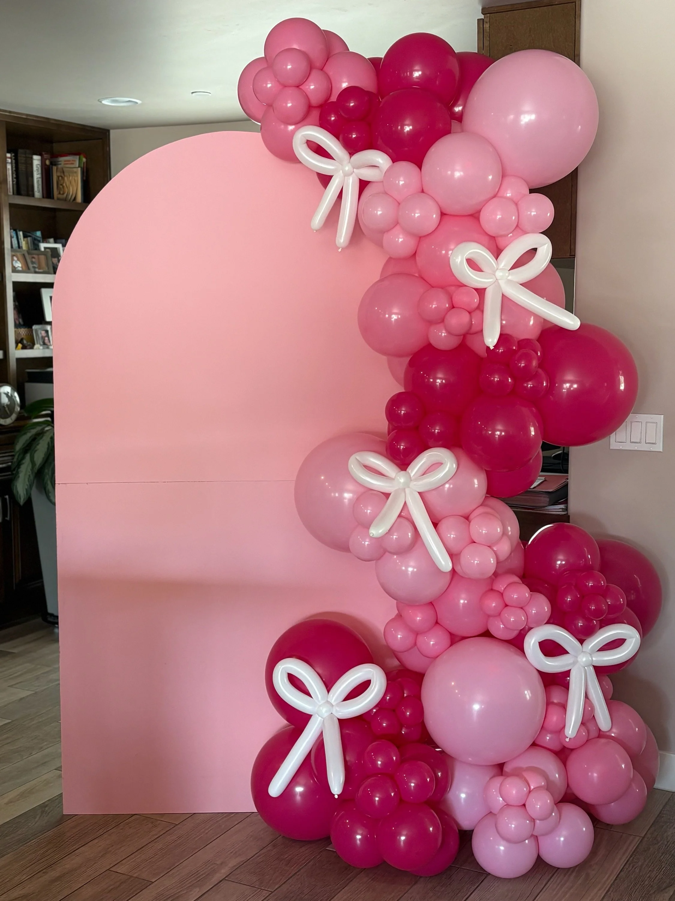 Pink backdrop decorated with clusters of pink, red, and white balloons arranged in a cascading manner, accented with white balloon bows.