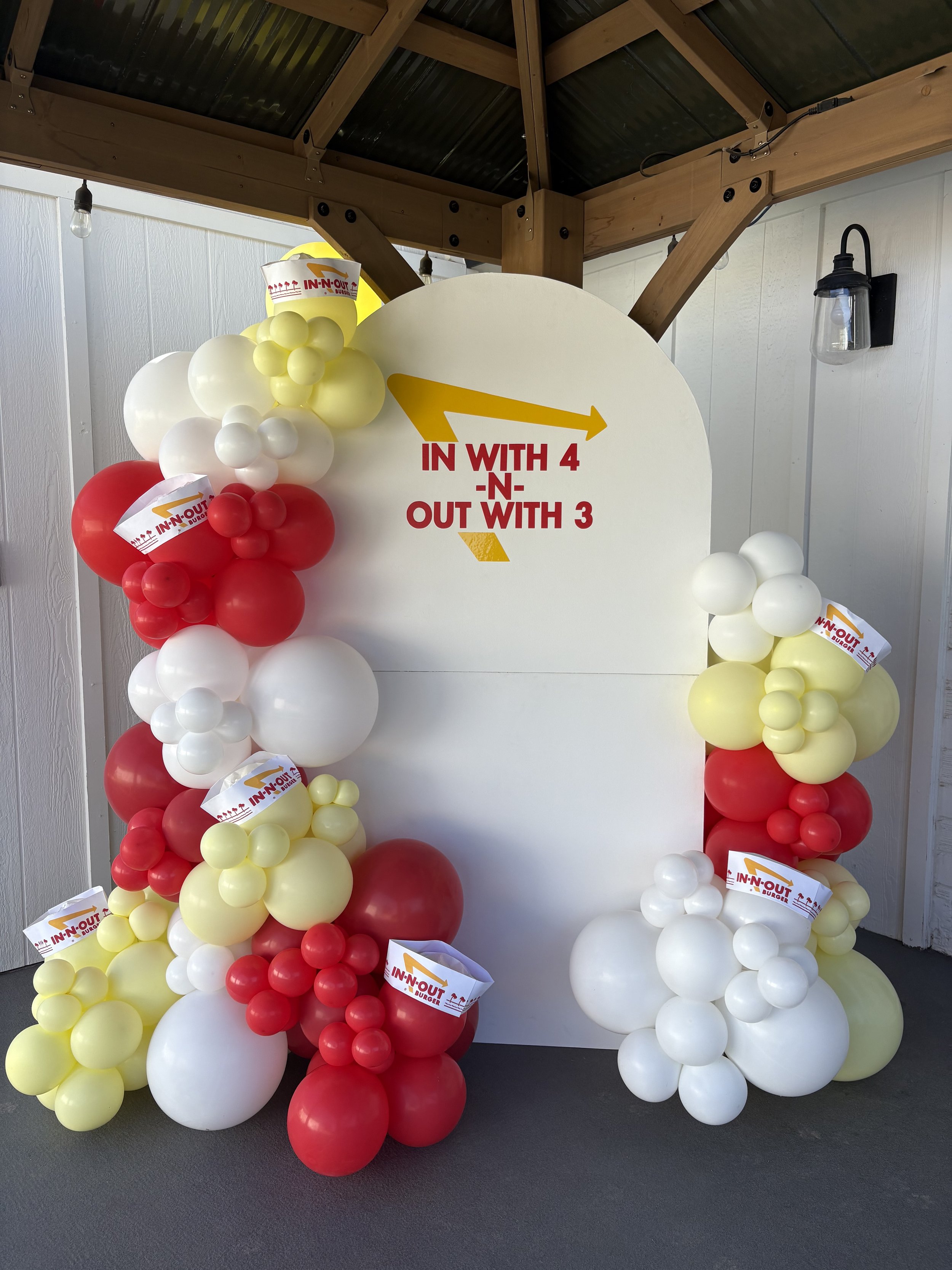 Decorative balloon display with yellow, white, and red balloons surrounding a white board that has an In-N-Out Burger sign and a directional arrow with the text 'In with 4 -N- Out with 3'.