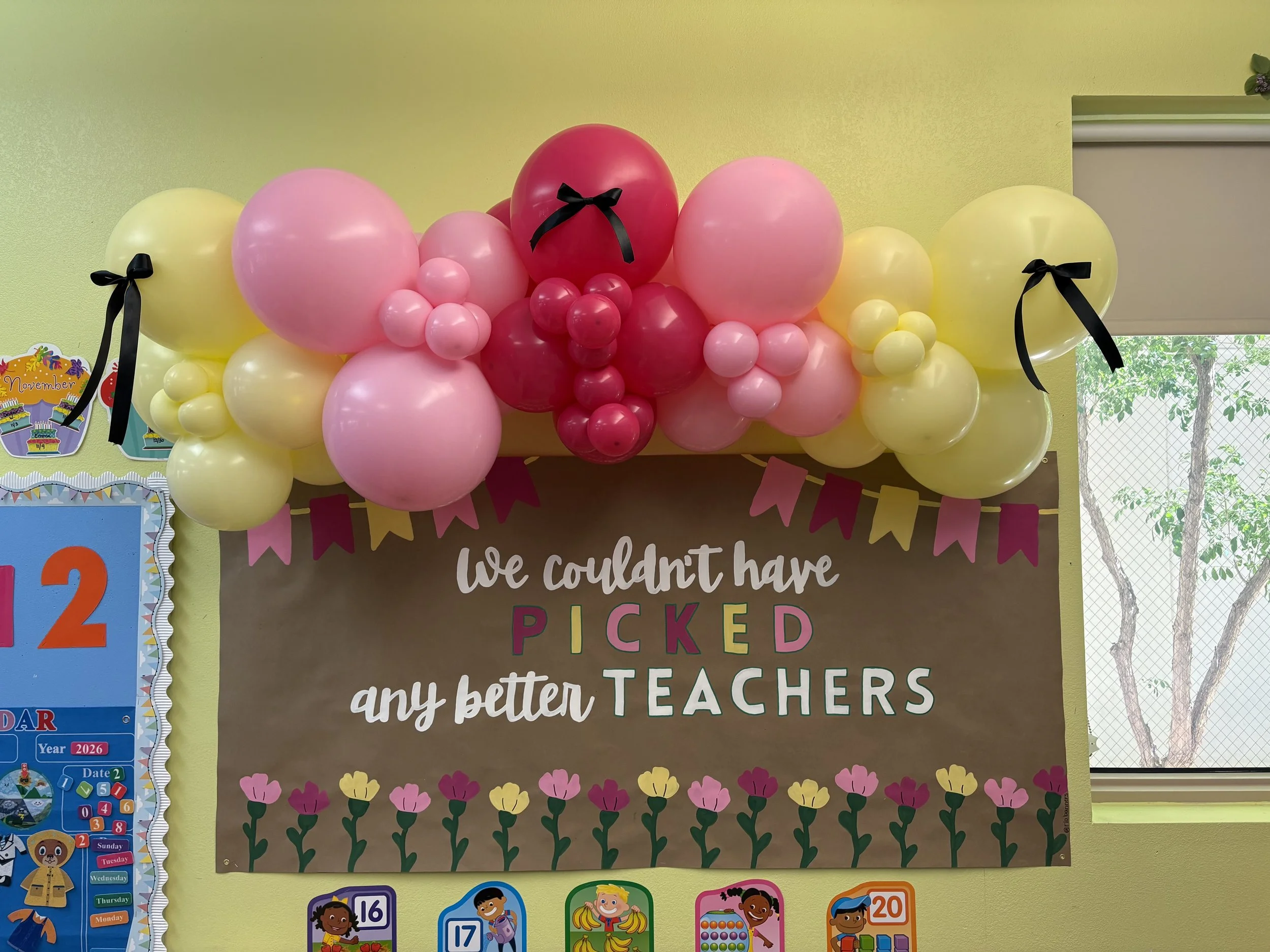 Decorative classroom bulletin board with pink, yellow, and red balloons arranged in an arch at the top, black ribbons tied around some balloons, a brown background poster with the phrase "We couldn't have picked any better teachers" in colorful lette