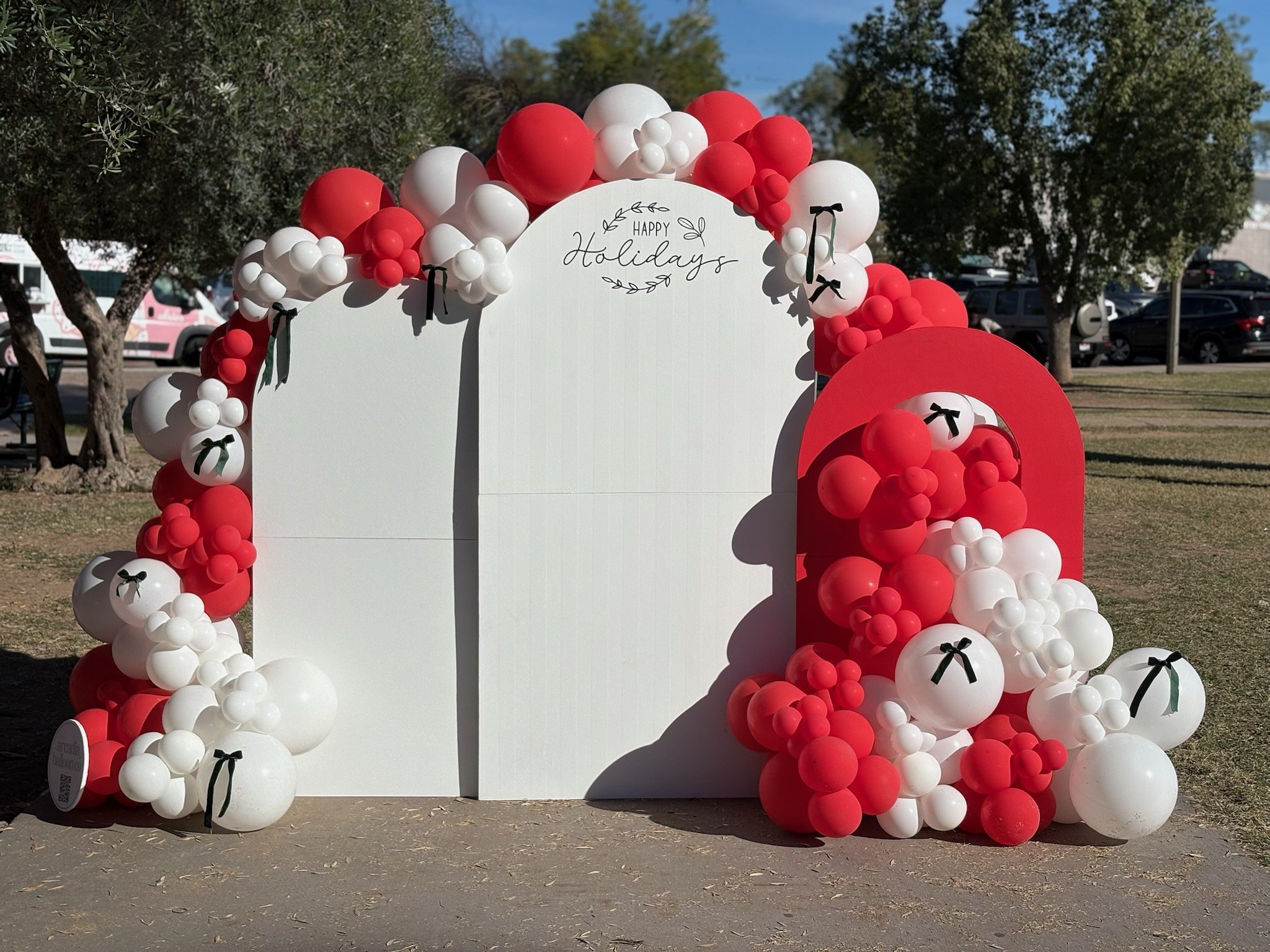 Decorative holiday display with a white and red balloon arch surrounding a white board that says 'Happy Holidays' and a red mailbox styled as a Christmas ornament with balloons.
