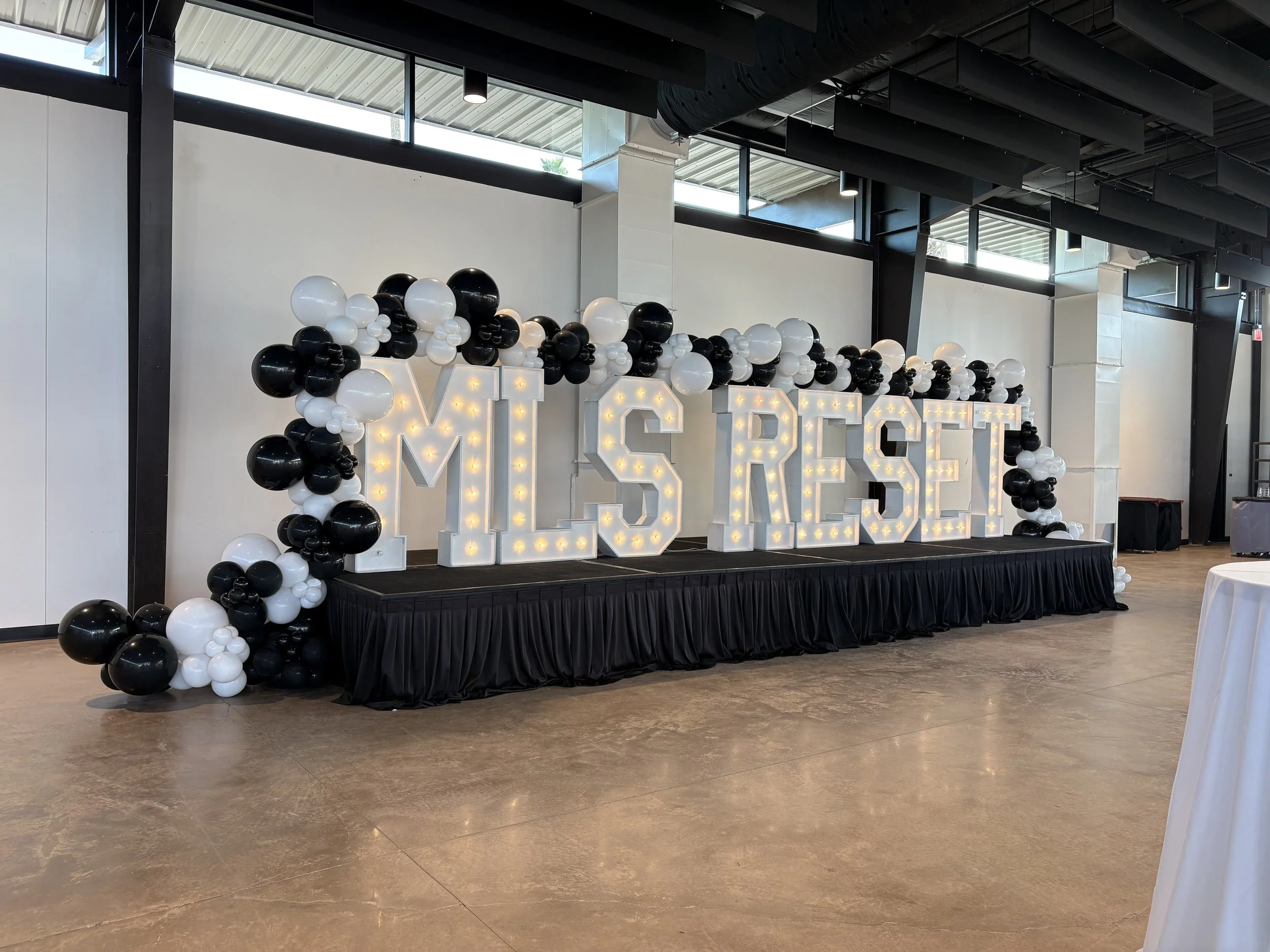 Large illuminated marquee letters spelling 'MLS RESF' on a black-draped stage, decorated with black and white balloon garlands, set in a spacious modern event hall.
