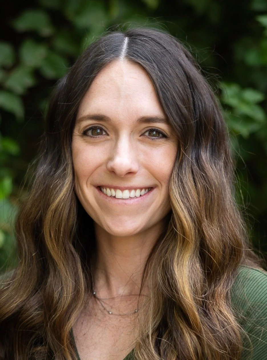 Close-up of a woman with long, wavy brown hair smiling outdoors with greenery in the background.