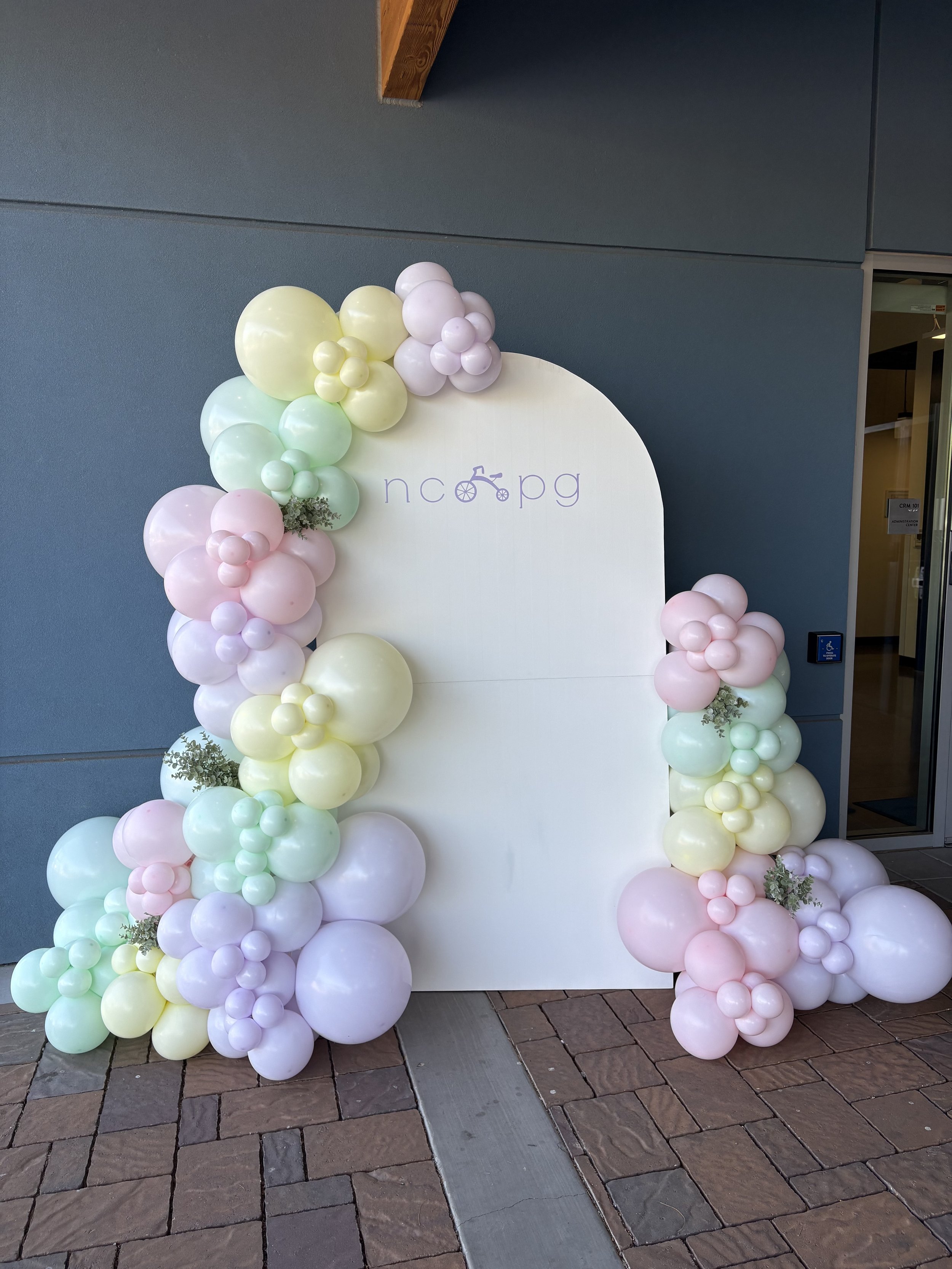 Decorative balloon arch with pastel colors surrounding a white backdrop that has the text 'ncapg' and a bicycle icon.