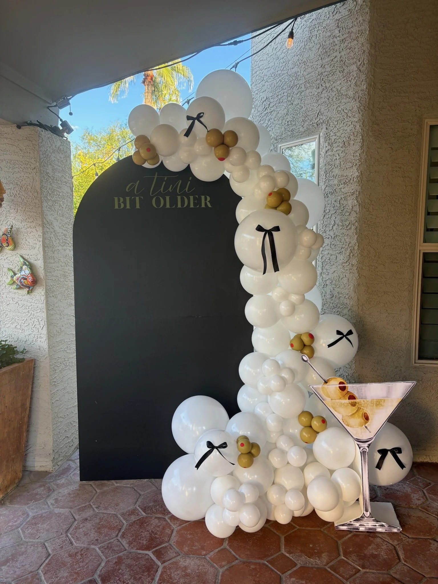 Decorative balloon arch with white, gold, and black balloons surrounding a black chalkboard sign that reads 'a tiny bit older' in cursive and block text. The arch includes balloon clusters with small black bows, and a large martini glass cutout with 