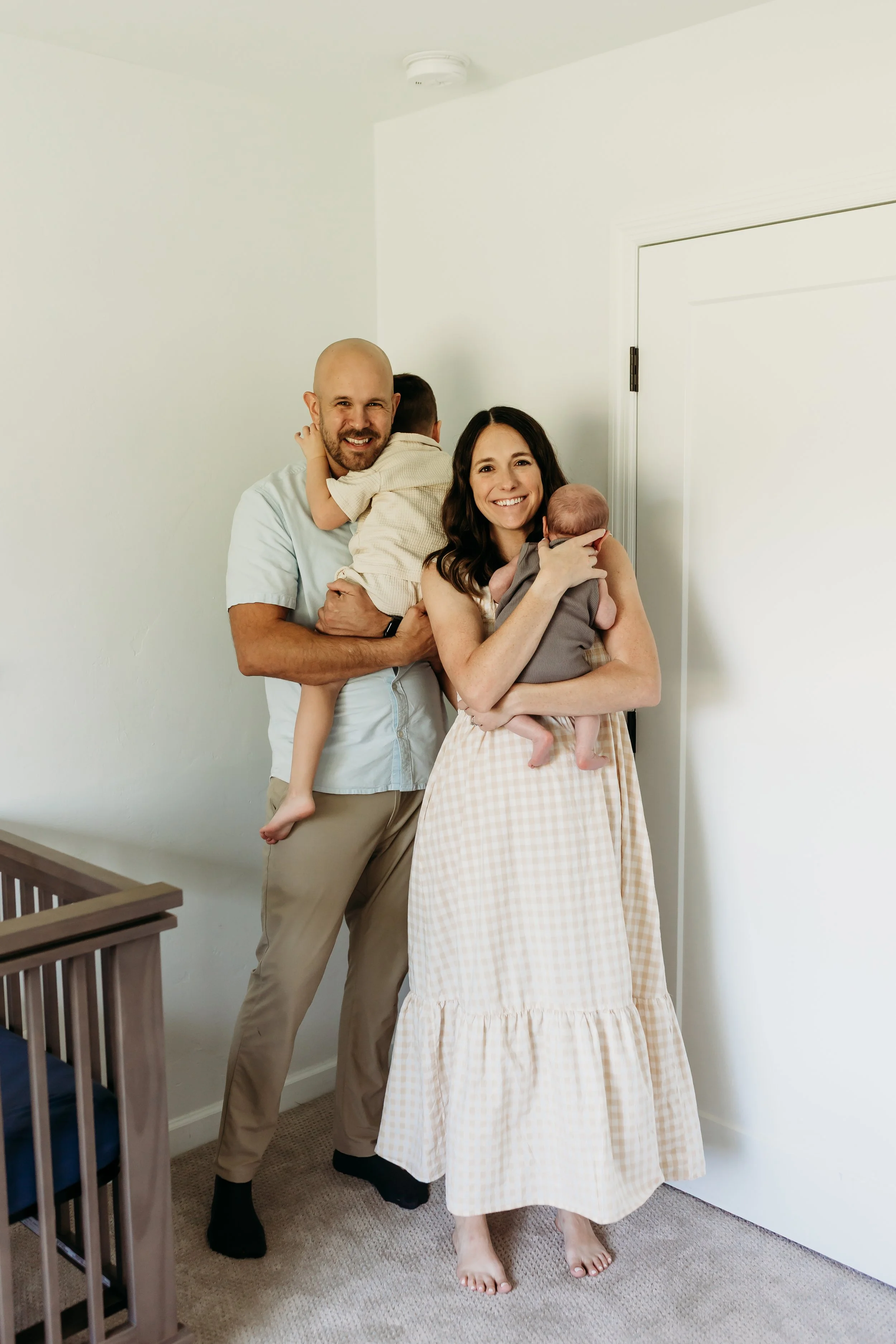 A smiling family of four, including a man, woman, and two children, standing indoors near a white door. The woman is holding a baby, and the man is holding an older child around his shoulders.