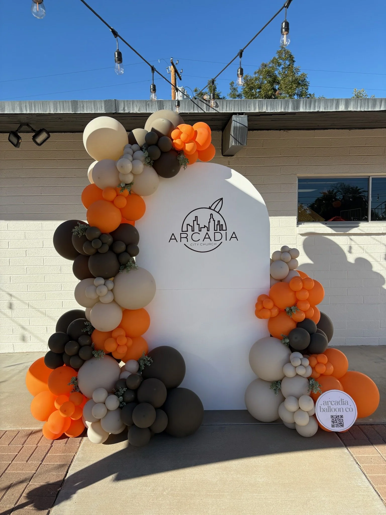 Balloon arch in orange, black, beige, and gray colors around a white backdrop with the Arcadia City Church logo, outdoors with string lights overhead.