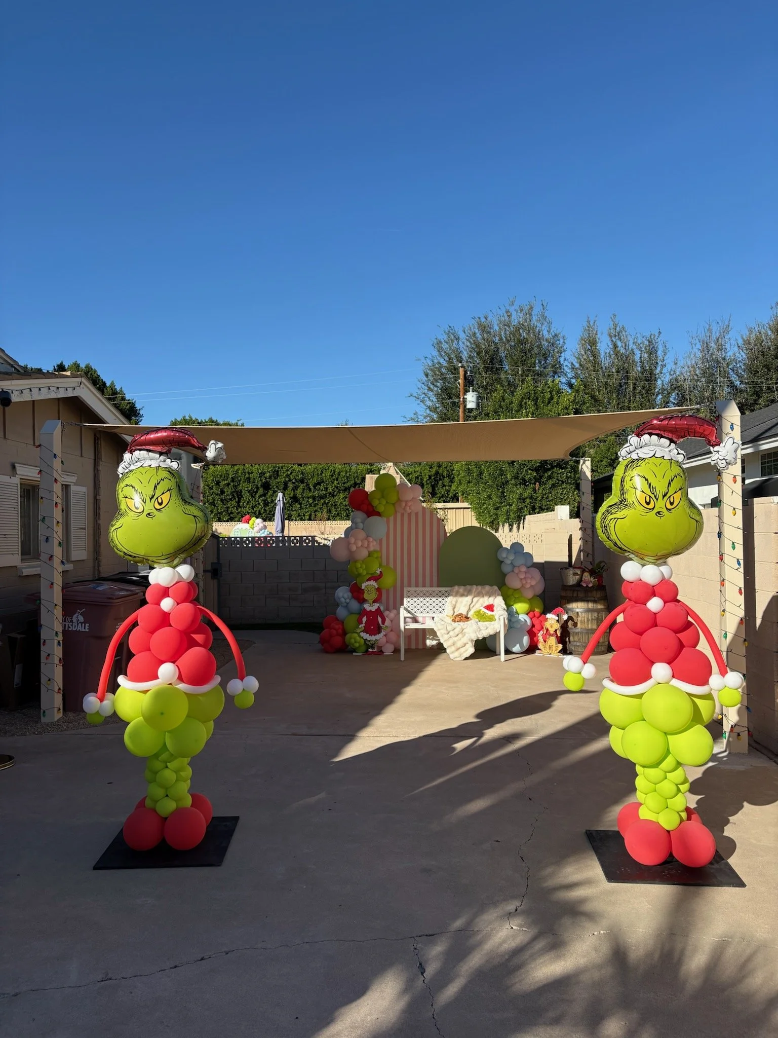 Colorful balloon decorations featuring Grinch faces on top of red and green balloon figures, set up for a Christmas celebration in a backyard with a desert concrete floor, a backdrop of trees, and a blue sky.
