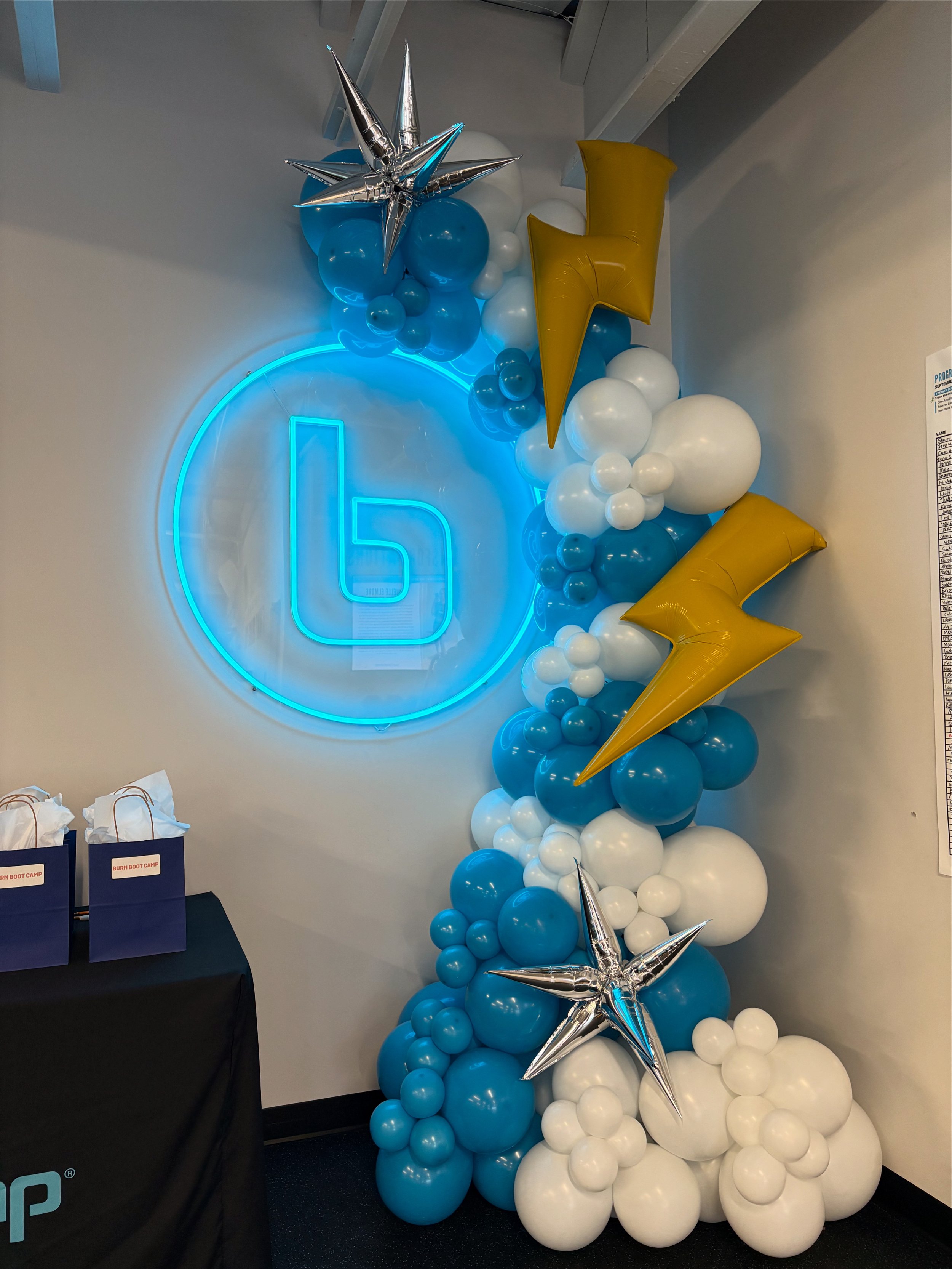 Decorative balloon arch with blue, white, and gold balloons, featuring silver star and gold lightning bolt-shaped balloons, illuminated by a blue neon logo on the wall.