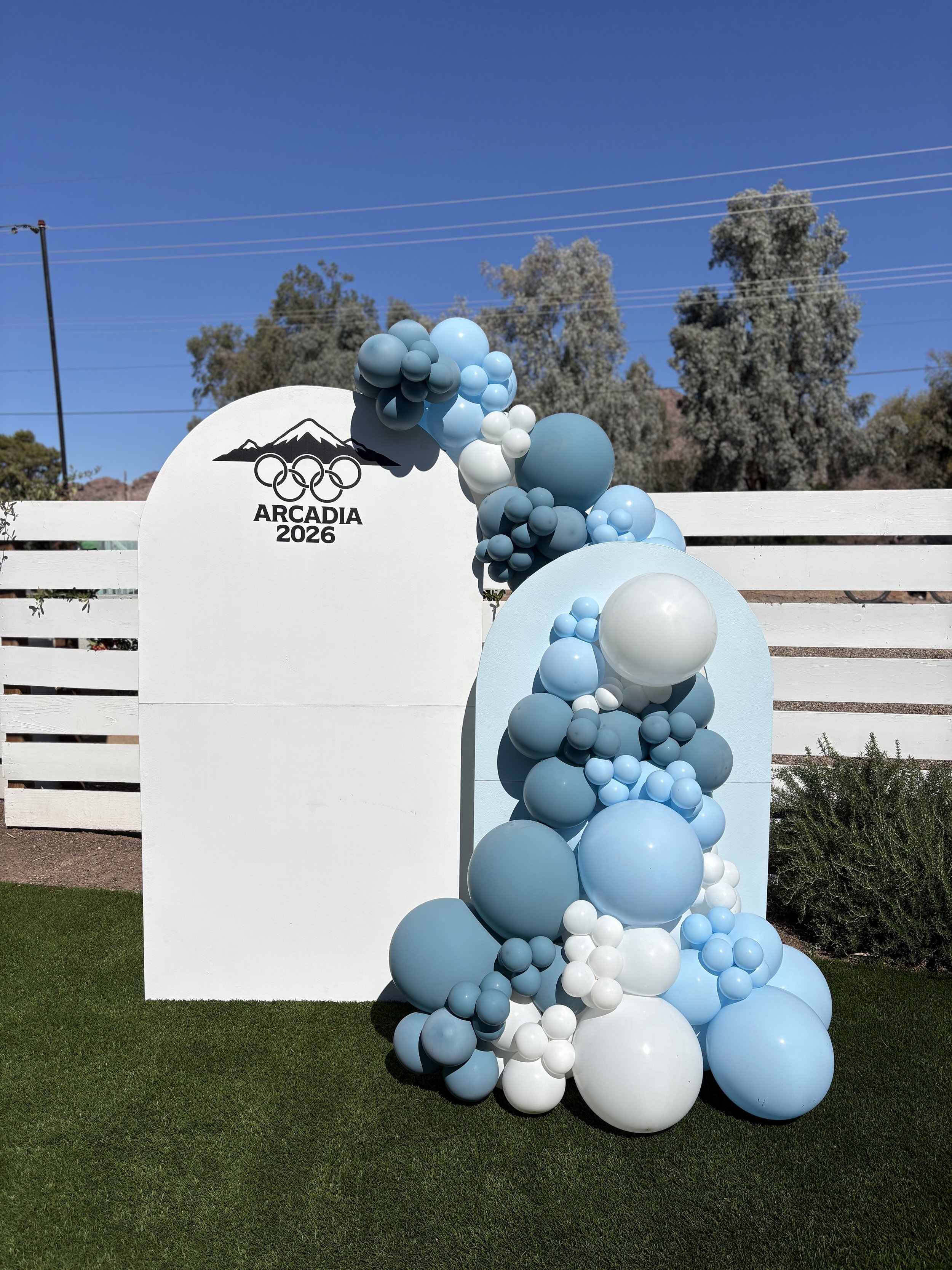 Decorative display with blue and white balloons, a white sign with the Olympic rings, mountain outline, and the text 'Arcadia 2026' in an outdoor setting with a white fence and trees in the background.