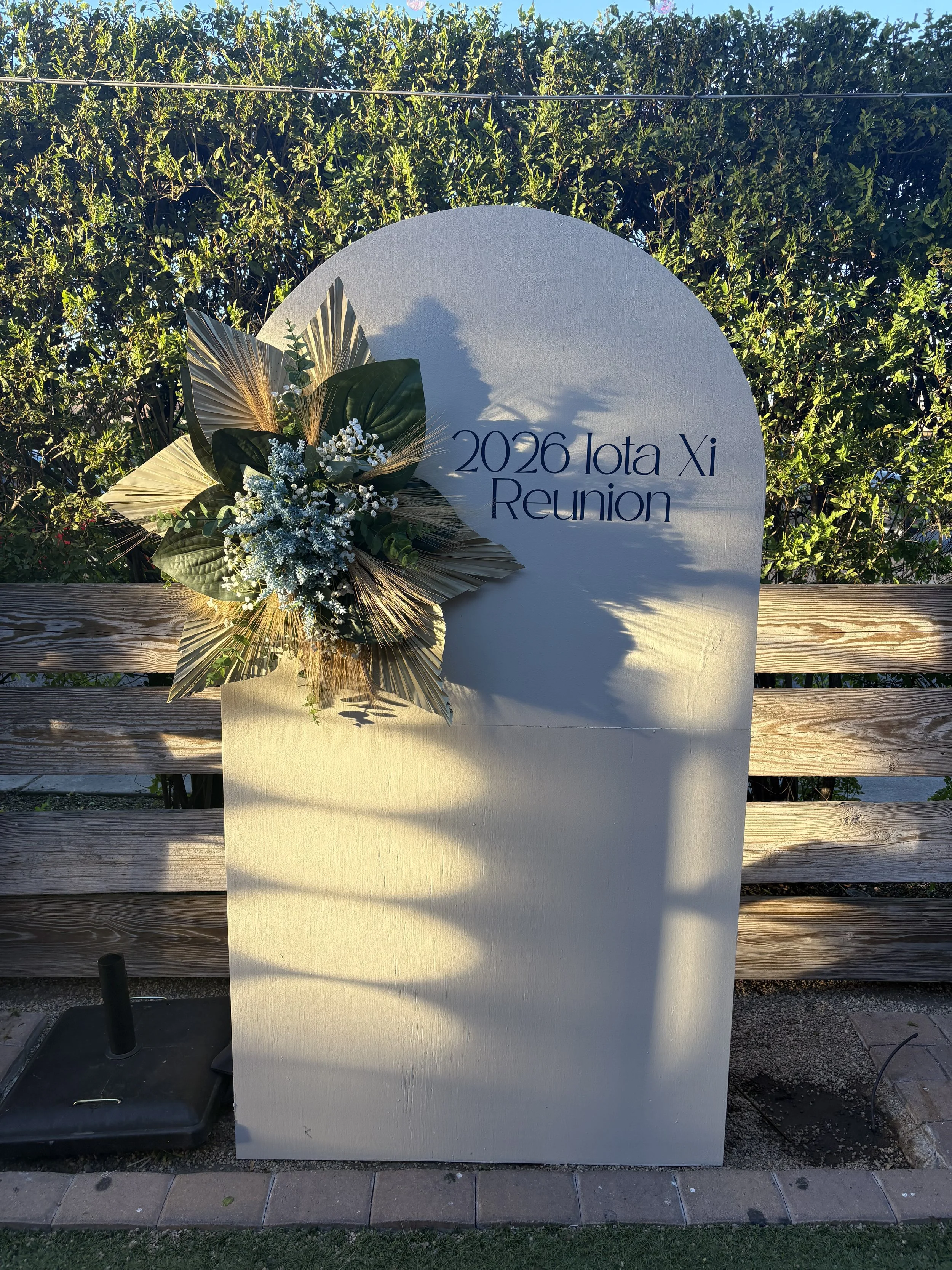 A sign with the text '2026 Iota Xi Reunion' decorated with a floral arrangement of greenery and white flowers, set outdoors against a wooden fence and green shrubbery.