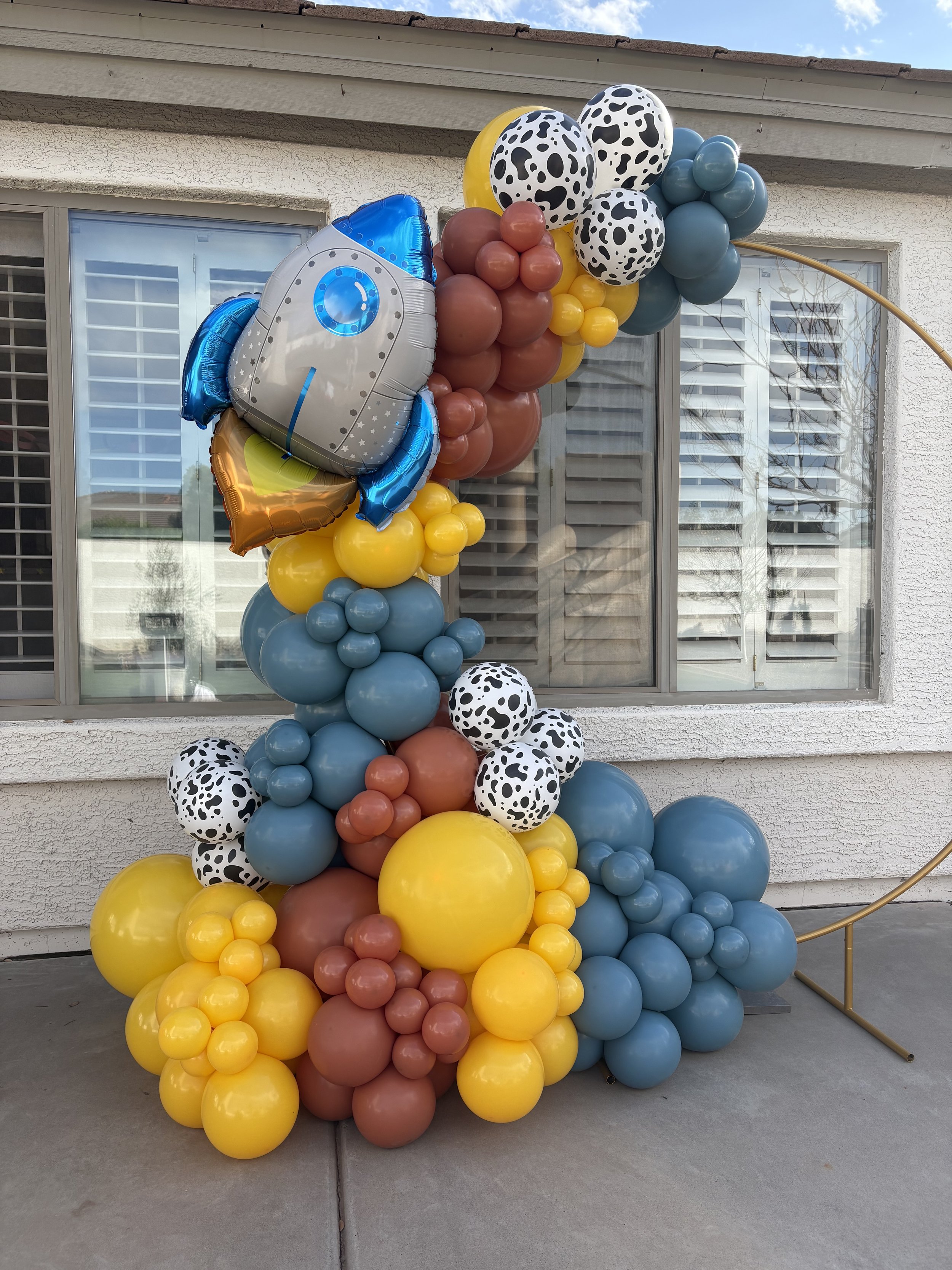 A balloon arrangement with a space-themed rocket ship and planets, featuring blue, yellow, brown, and black-and-white spotted balloons, set outside against a house wall.
