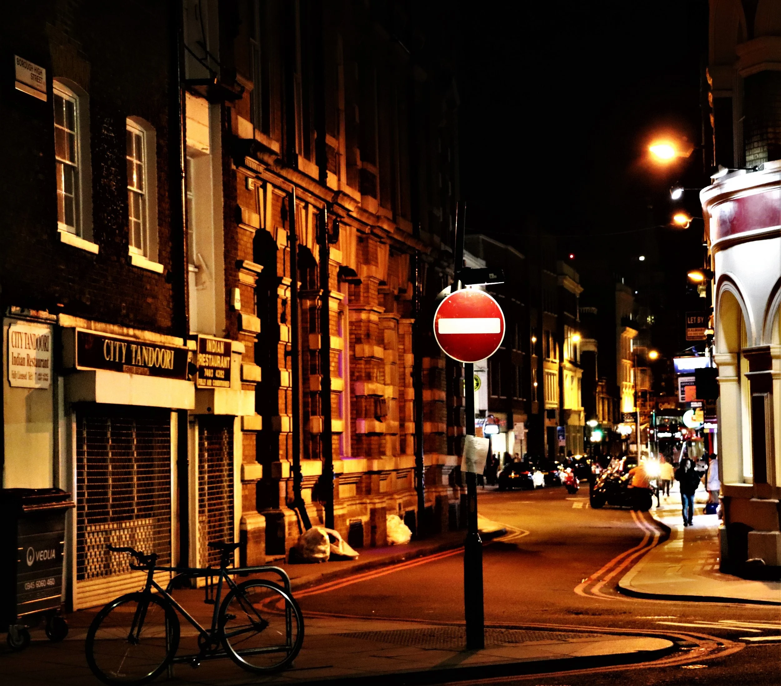 Nighttime city street with illuminated buildings, parked cars, a bicycle, a no-entry sign, streetlights, and people walking.