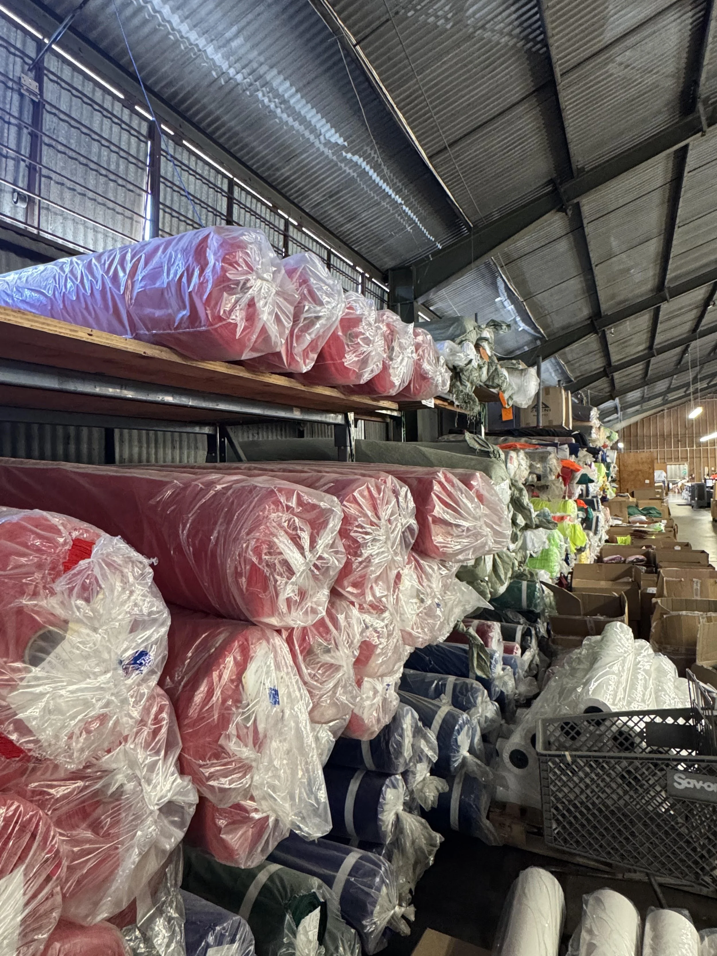 Rows of large plastic bags filled with colorful textiles stored on metal shelves inside a warehouse.