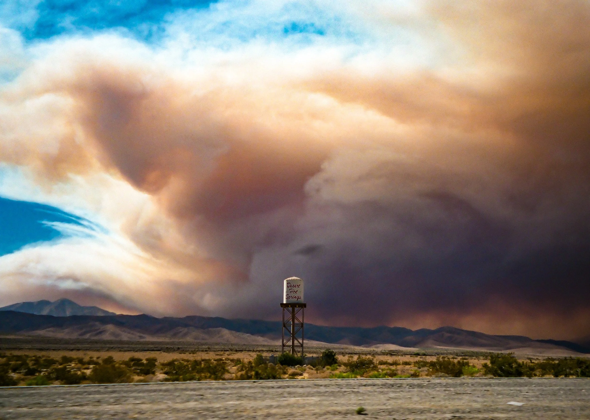 A large sky with scattered clouds during either sunrise or sunset over a mountainous desert landscape. In the foreground, there's a sign that reads 'Desert Hot Springs' on a tall metal structure.