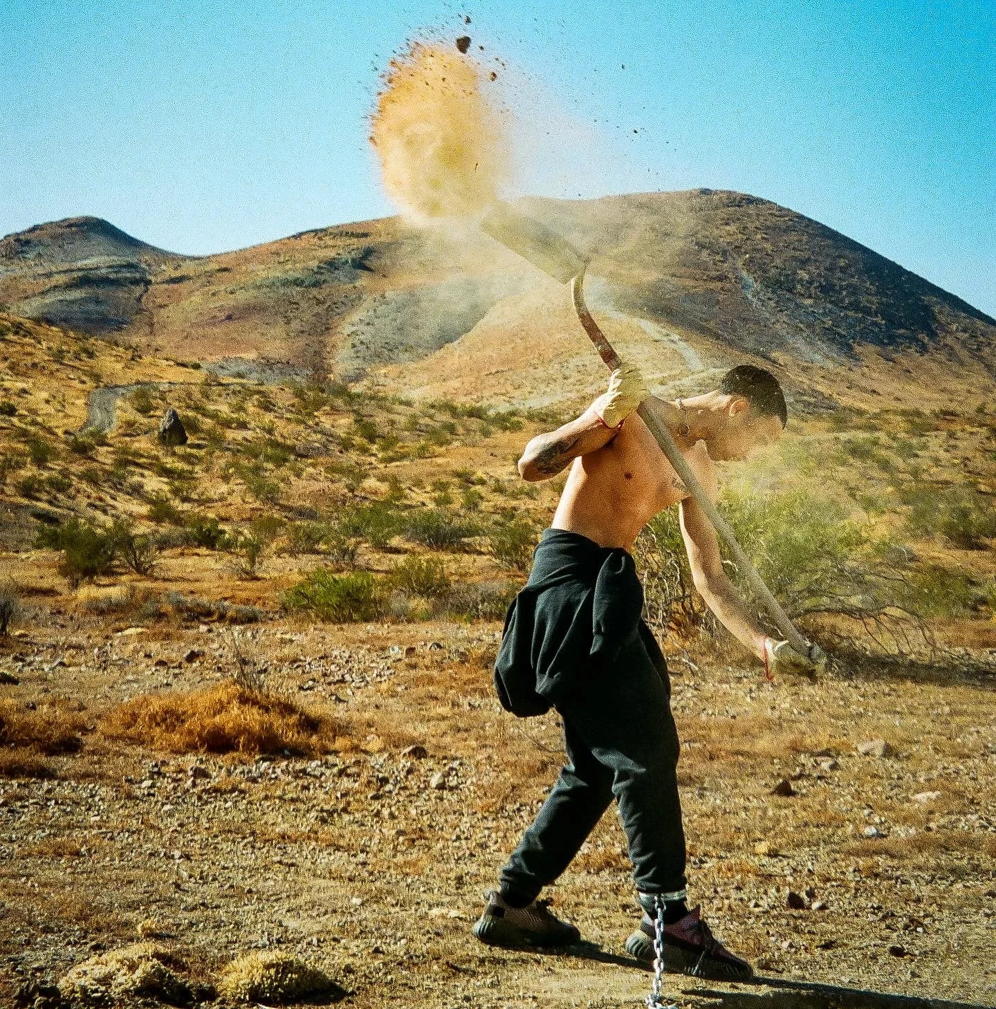 A shirtless man with black hair and tattoos is chained to the ground in a desert landscape. He is holding a large flaming sword above his head, and sparks or ash are flying from it. The background features hills or mountains and sparse vegetation.