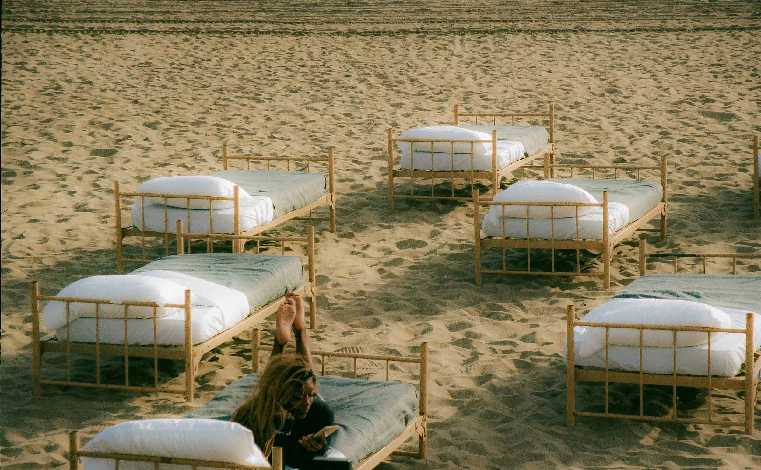 Multiple empty wooden beds with white mattresses and pillows arranged on the sandy beach, with a woman sitting on one reading a book.