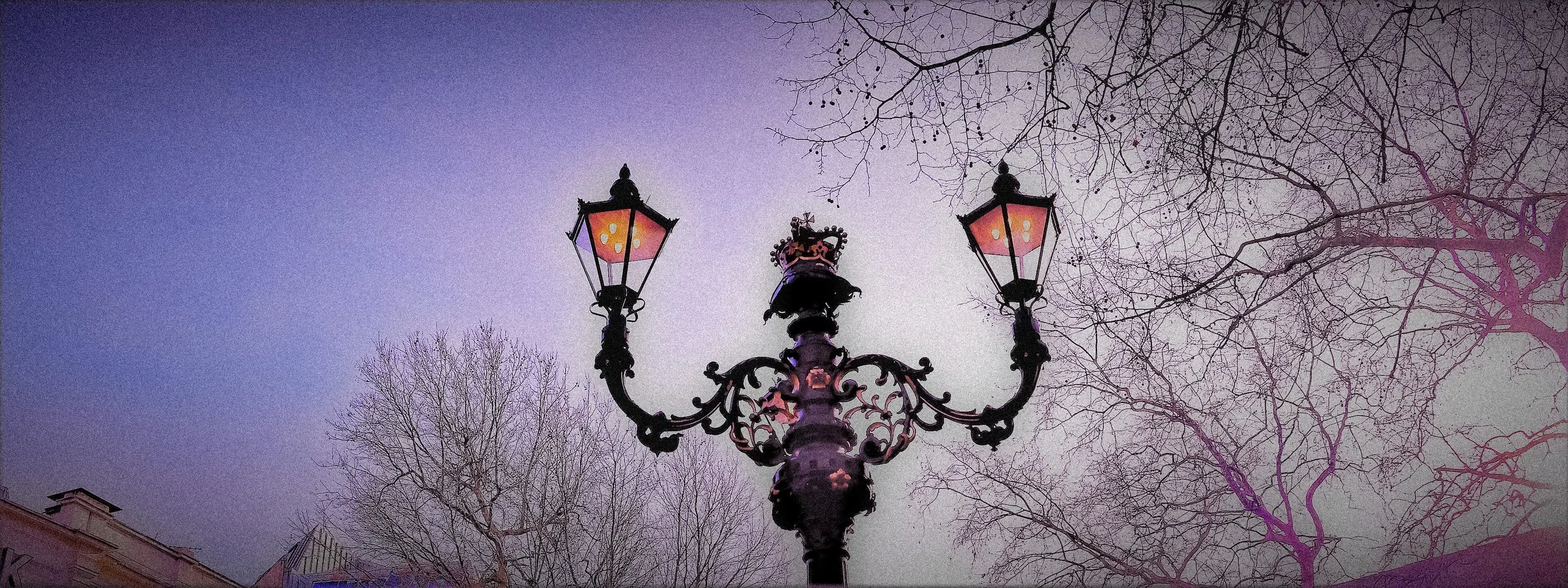 A vintage street lamp with three lanterns, turned on, against a dusk sky. Bare tree branches are visible in the background.