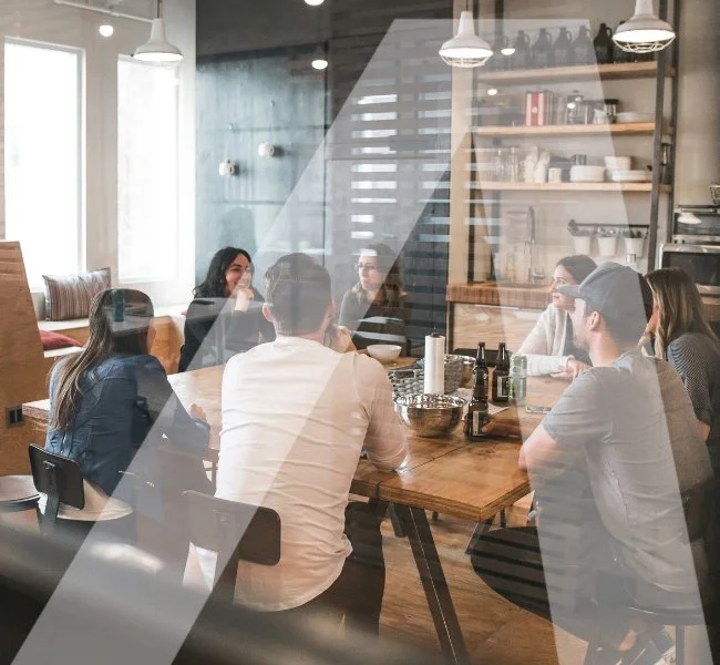 Group of people having a meeting in a modern, well-lit cafe or restaurant with wooden furniture and open shelving on the wall.