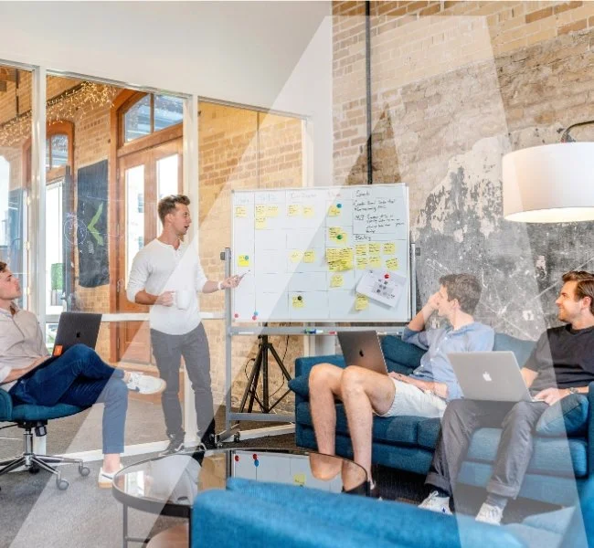 A group of four people in a modern office or meeting room, with one person standing and presenting near a whiteboard filled with sticky notes, while the others are seated, some with laptops, listening attentively.