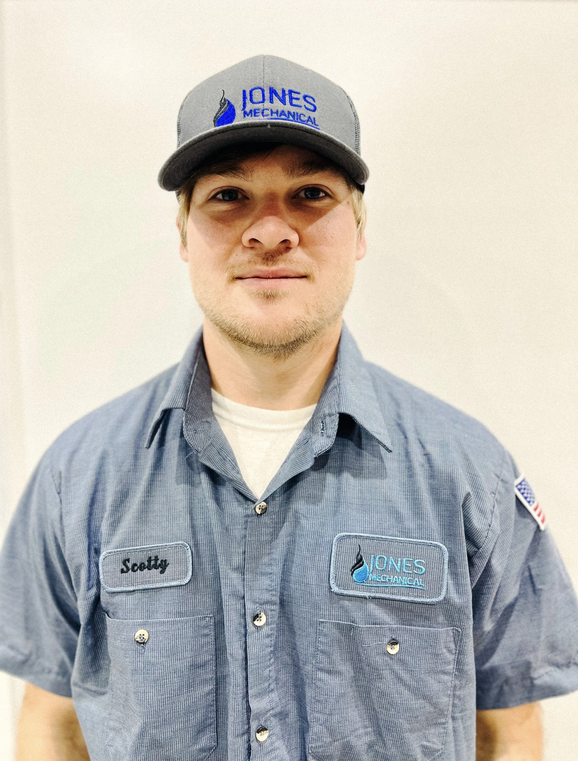 A young man with light skin, blond hair, and a slight beard standing against a white background. He's wearing a gray work uniform with a name tag that reads 'Scotty' and a logo badge of 'Jones Mechanical.' He's also wearing a gray cap with the same company logo.