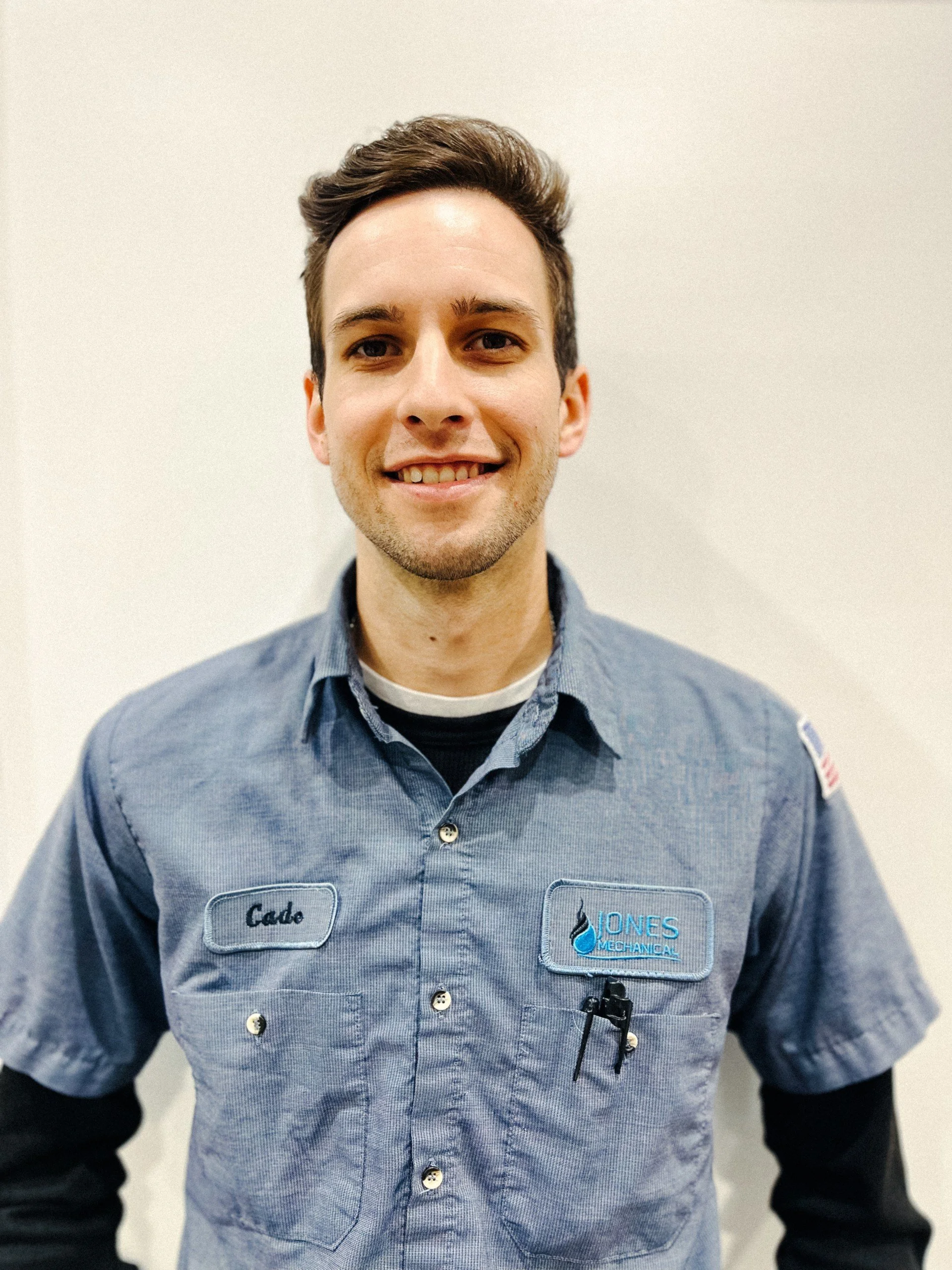 A young man smiling, wearing a blue work shirt with a name tag that says Gade, and a logo that says Jones Mechanical.