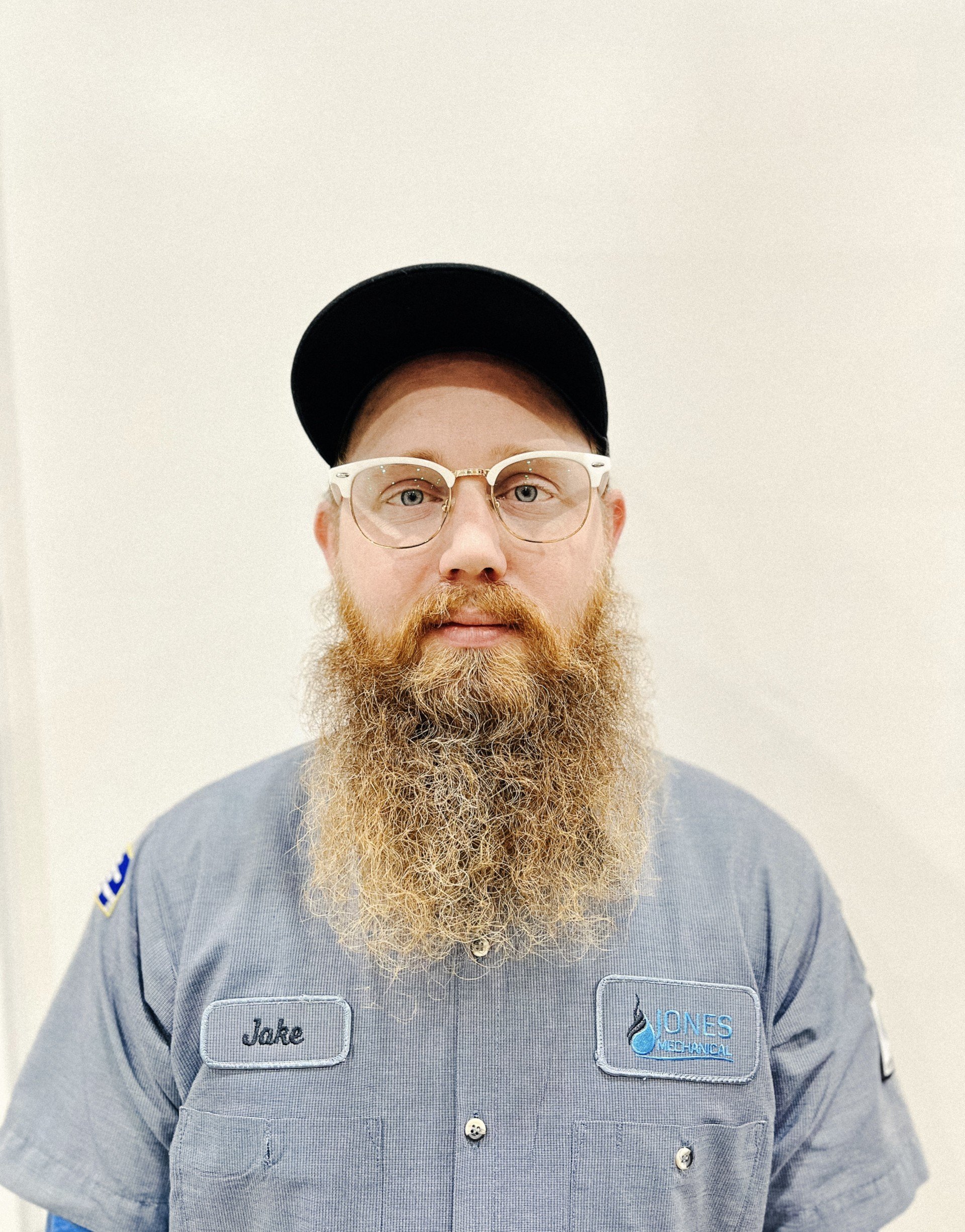 A man with a long beard, glasses, and a black cap wearing a gray work shirt with a name tag that says 'Jake' and a patch from Jones Mechanical.