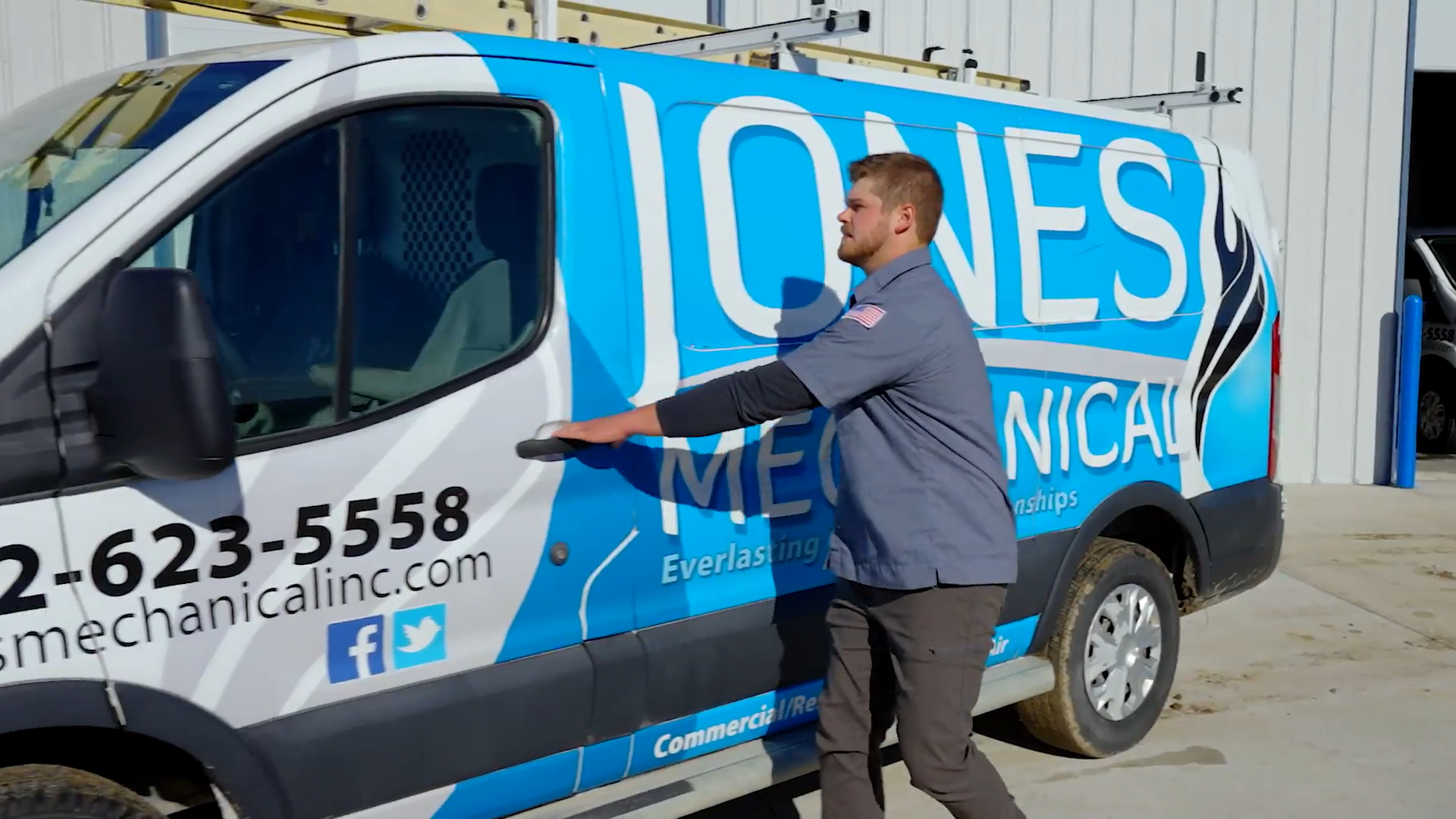 A technician closing the door of a blue and white service van with the company name 'Jones Mechanical' and contact information printed on the side.