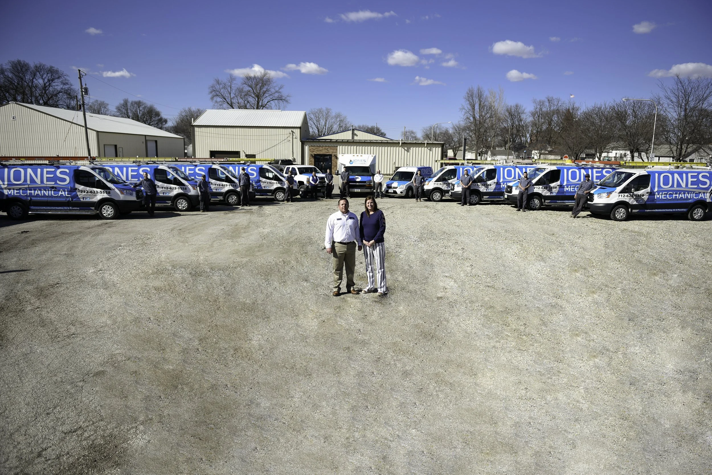 Two people standing in the center of a gravel lot with a line of company vehicles and employees behind them, industrial buildings and leafless trees in the background, under a partly cloudy blue sky.