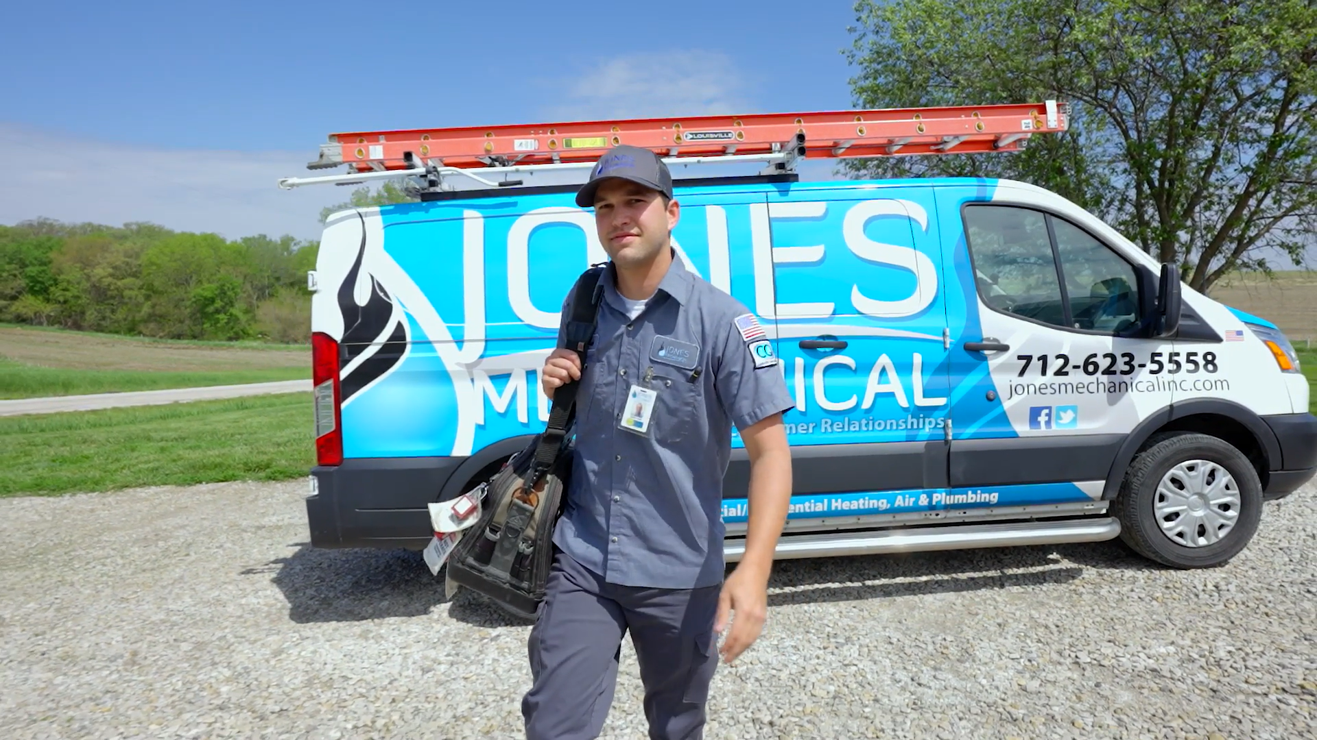 A man in gray work uniform with a backpack stands in front of a blue Jones Mechanical service van on a gravel area with green grass and trees in the background.
