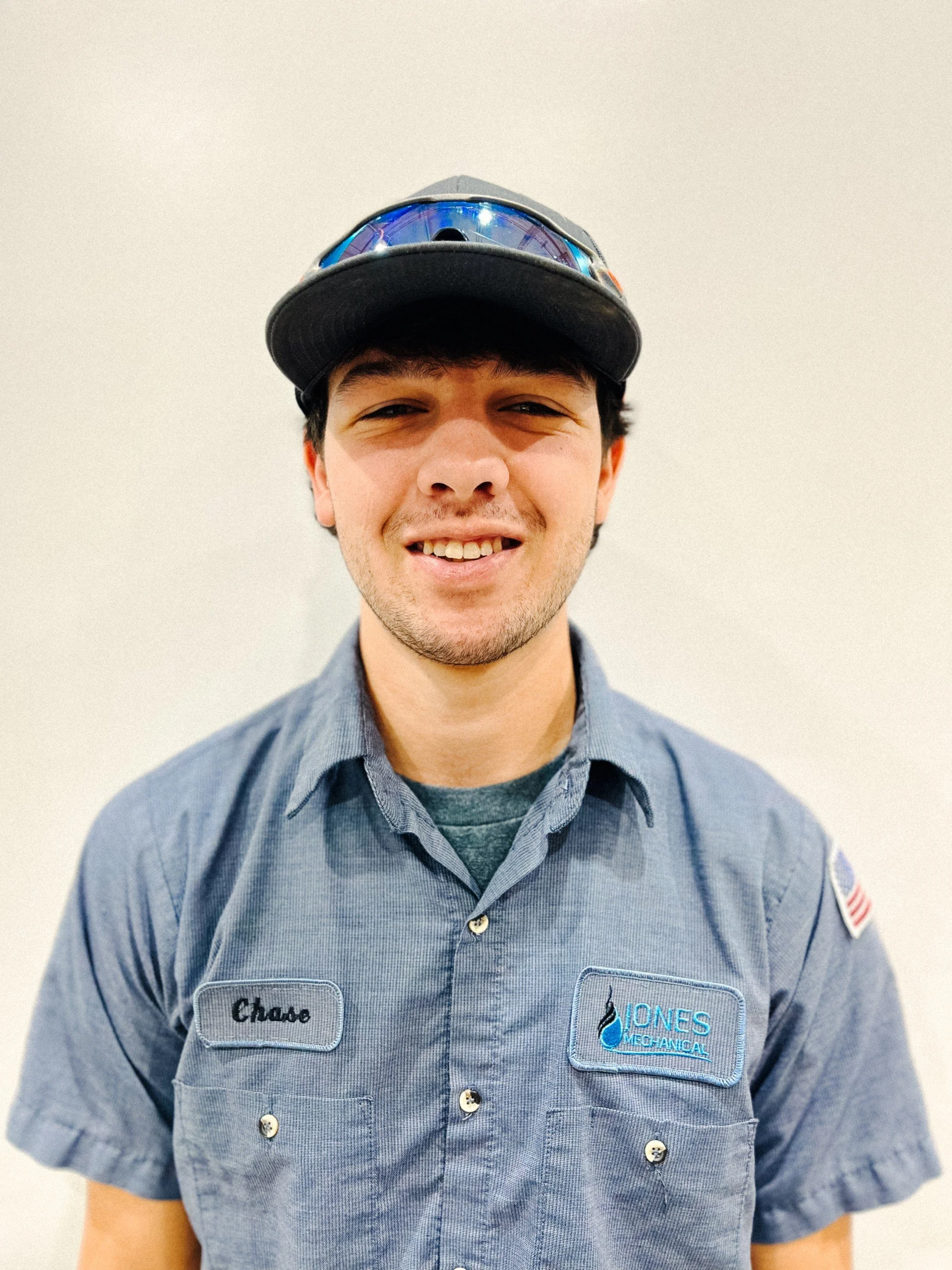 A young man in a mechanic's uniform with a name tag that says 'Chase' and a patch for 'Jones Mechanical', wearing a cap and sunglasses on top, smiling at the camera.