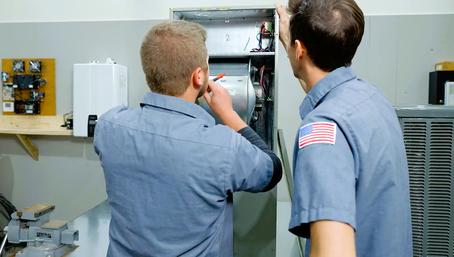 Two technicians in blue uniforms repairing or maintaining an electrical or HVAC unit in a workshop or maintenance room.