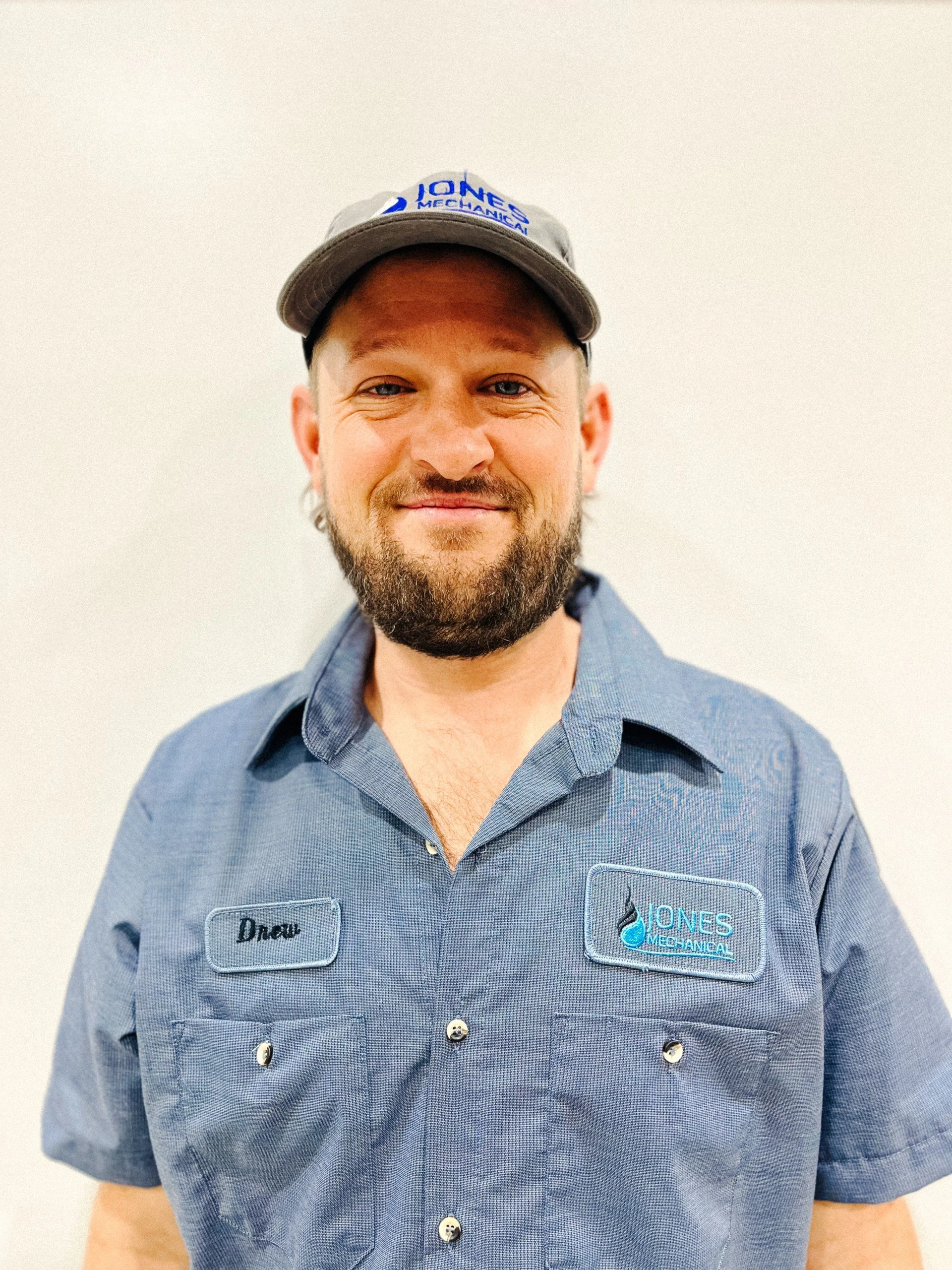 A smiling man in a blue work shirt and cap with a beard, standing in front of a plain white background.