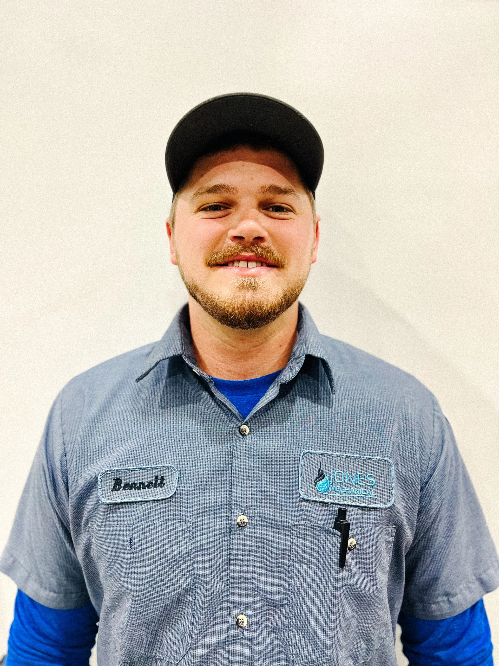 A smiling young male employee in a uniform with a name tag reading 'Bennett' and 'Jones Mechanical,' standing against a plain white wall.