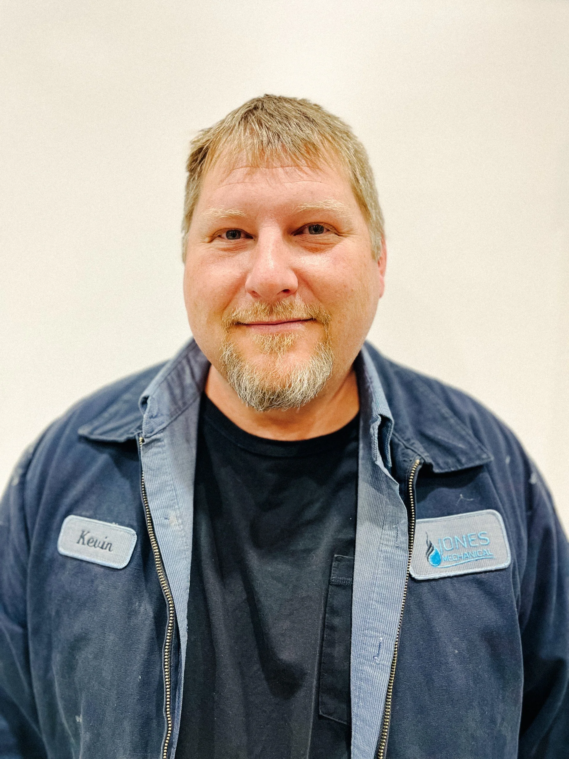 A man with light-colored hair and a beard, wearing a navy uniform with a nametag that says 'Kevin' and a patch reading 'Jones Mechanical,' standing against a plain white background.