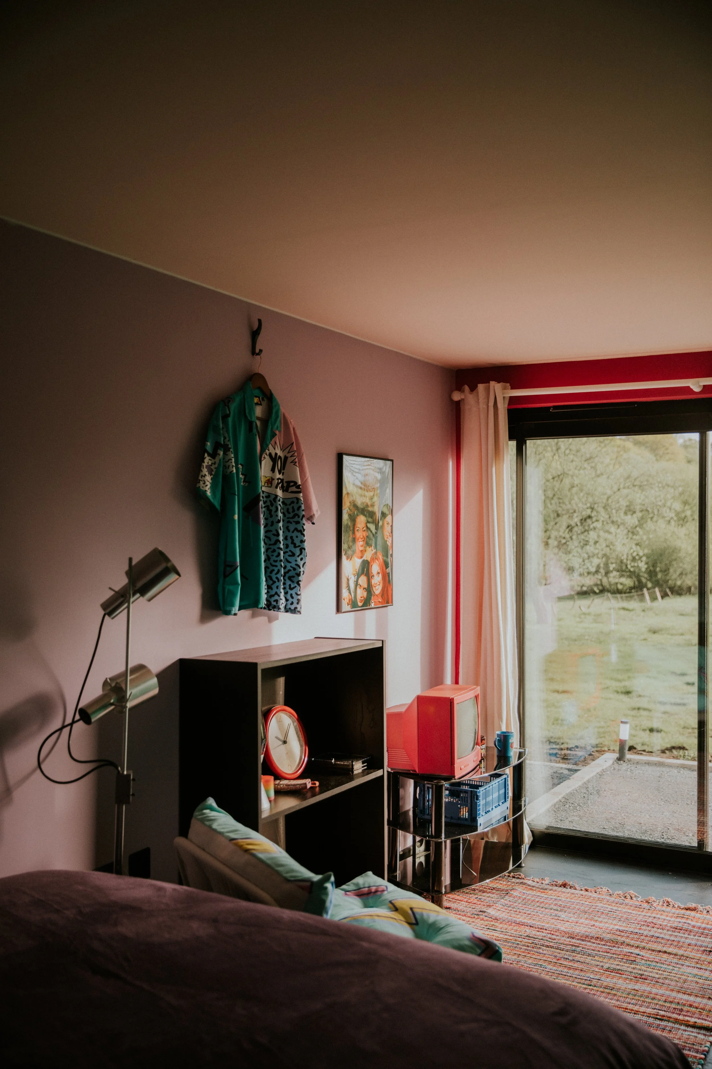 A cozy living room with a pink wall, a black shelf with a clock, a vintage pink TV, a small table with a black crate, and a large window with white curtains showing a green outdoor landscape.