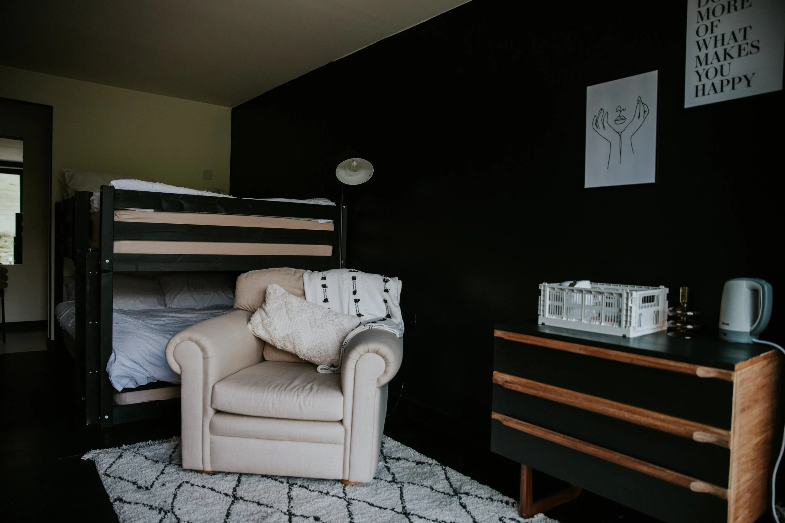 A cozy bedroom corner featuring a bunk bed with white bedding, a cream-colored armchair with a throw blanket, a black wall with framed artwork, a wooden dresser holding a white dish rack, a kettle, an air fryer, and a lamp, with a window on the left 
