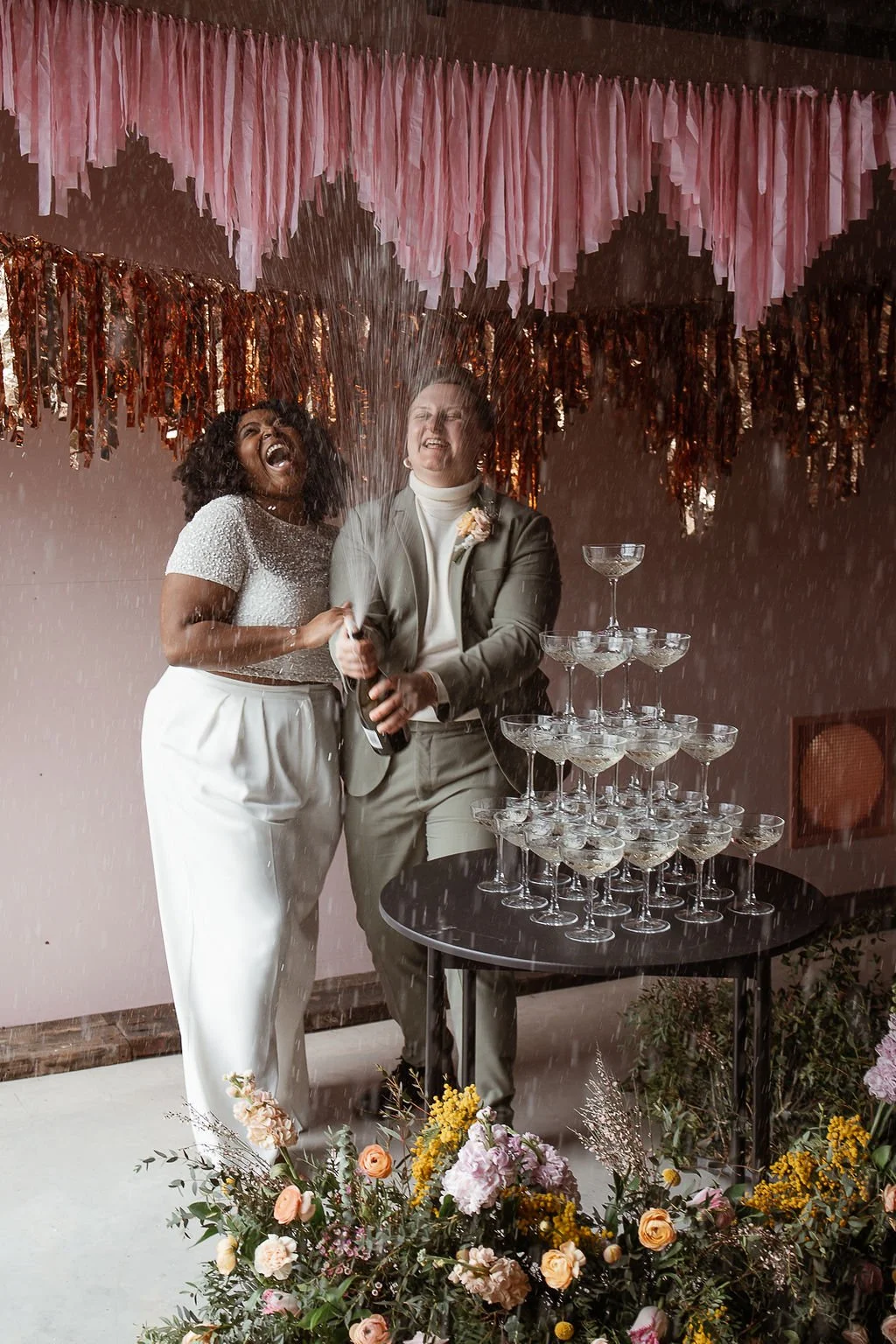 Two people, one in white pants and a beaded top and the other in a suit, celebrating with a champagne bottle and spray, standing next to a pyramid of champagne glasses on a table, with pink and orange decorations overhead and flowers in the foreground.