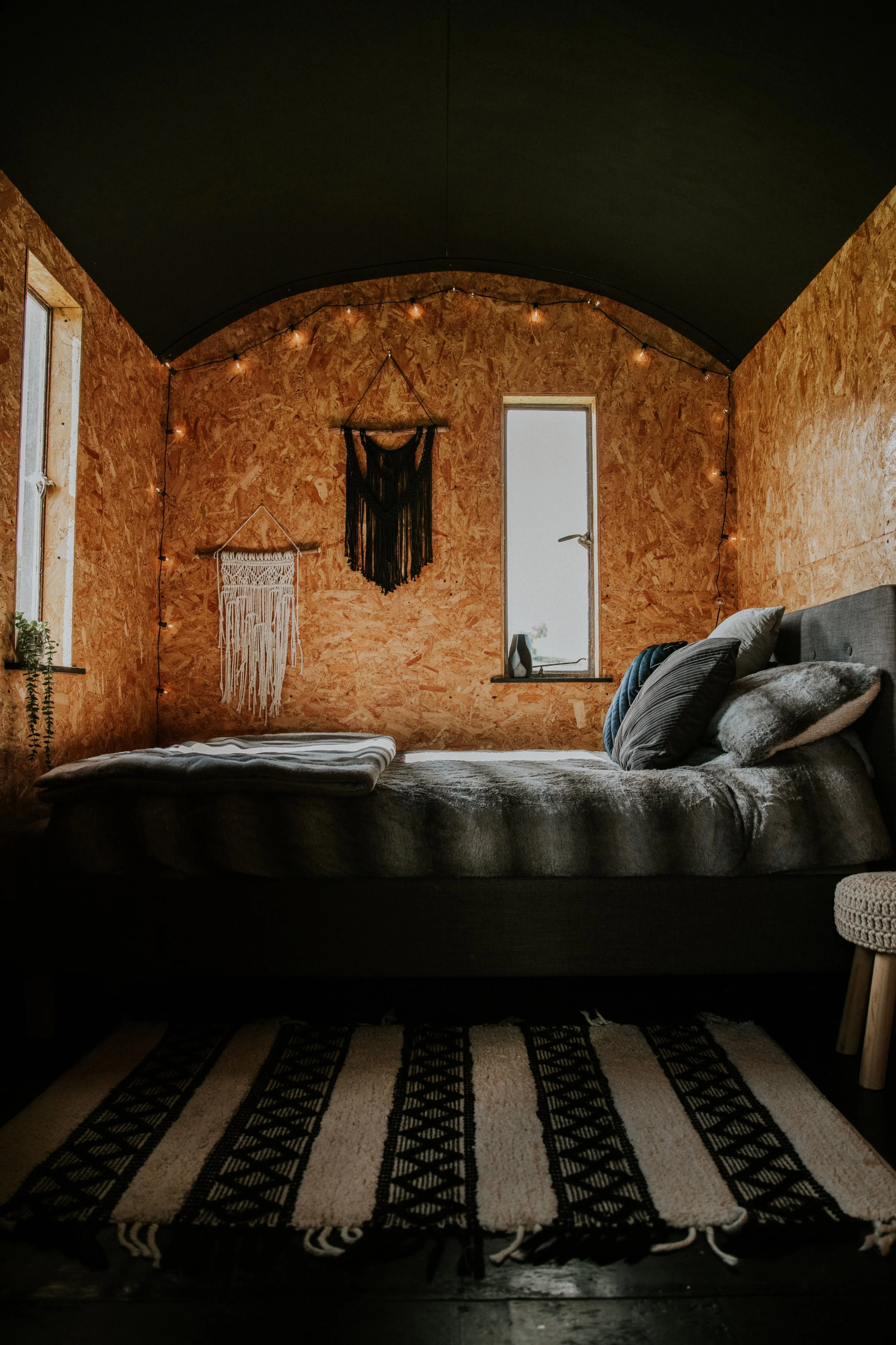 A cozy bedroom with an unfinished wooden wall, black ceiling, string lights, and two wall hangings. There is a bed with pillows and a blanket, and a black and white patterned rug on the floor.