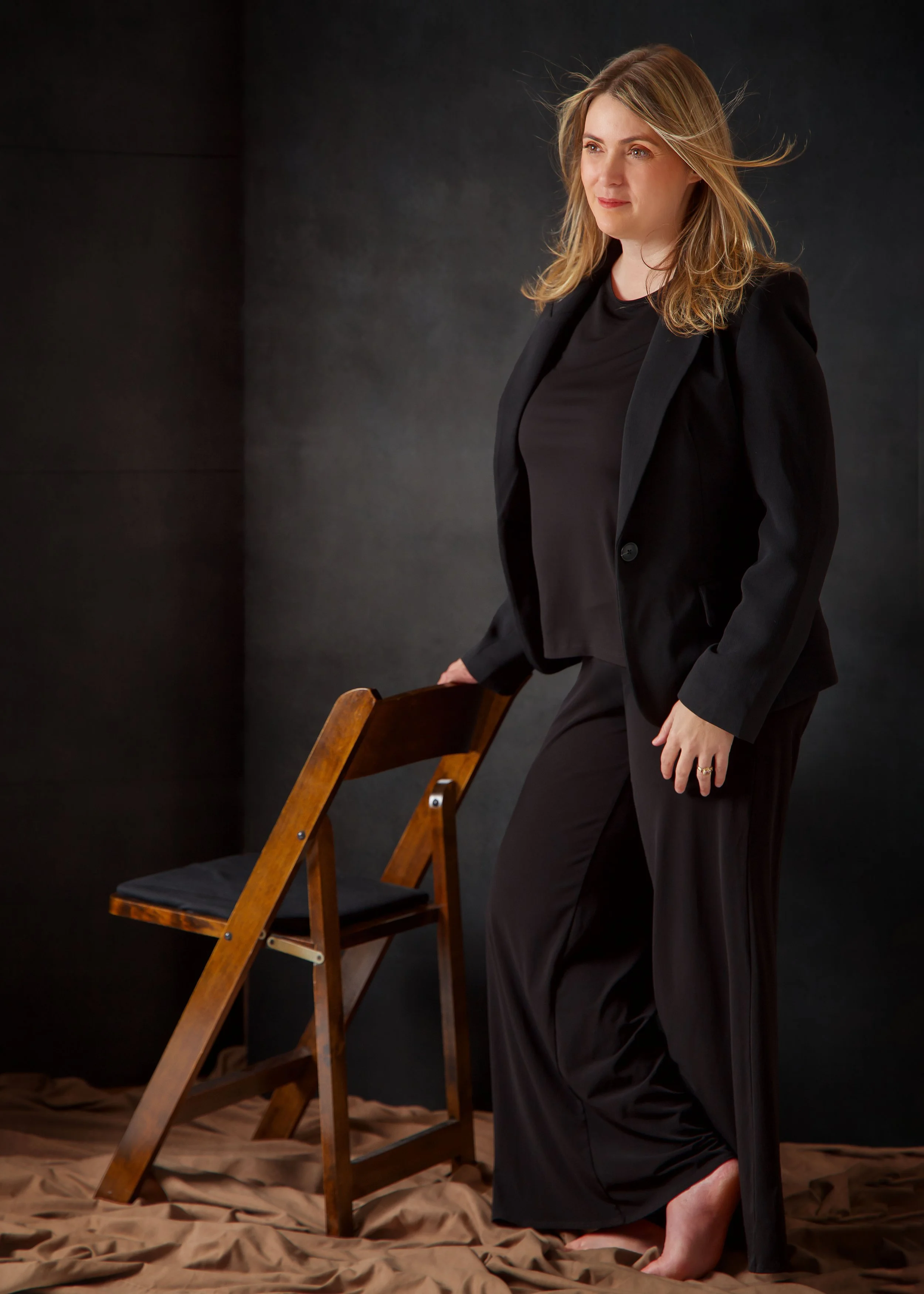 A woman in a black blazer and black pants standing next to a wooden chair with a black cushion, against a dark background.